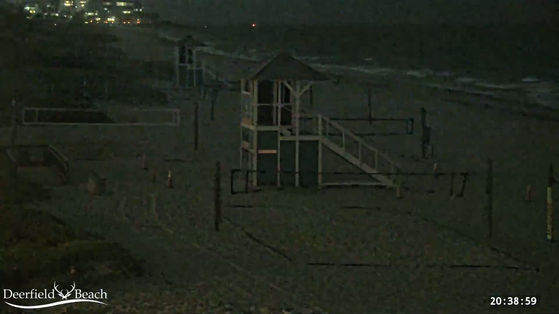 A dark, nighttime beach scene shows a lifeguard stand, volleyball nets, and a single person walking near the water under cloudy skies.