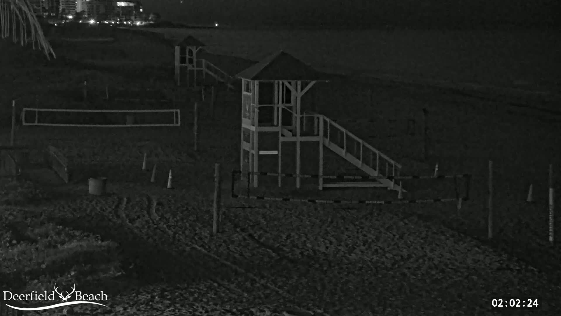 A nighttime, low-light image shows a sandy beach with volleyball nets, cones, and lifeguard stands.