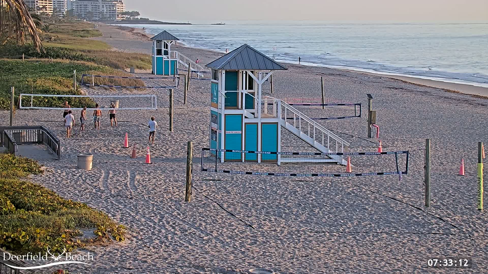 A sandy beach with several volleyball nets, lifeguard stands, and a group of people playing volleyball under a mostly clear sky.