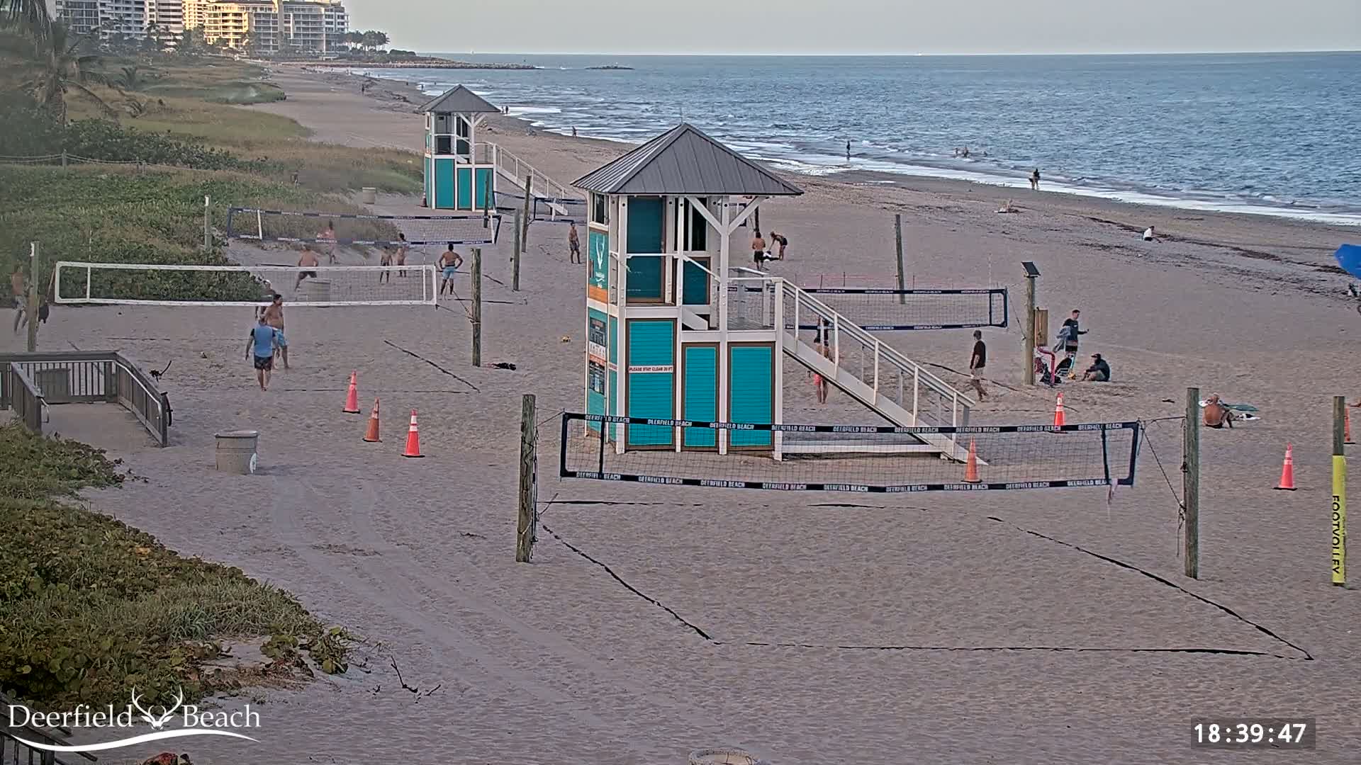 A sandy beach with several volleyball courts, lifeguard stands, and people playing volleyball under a partly cloudy sky.