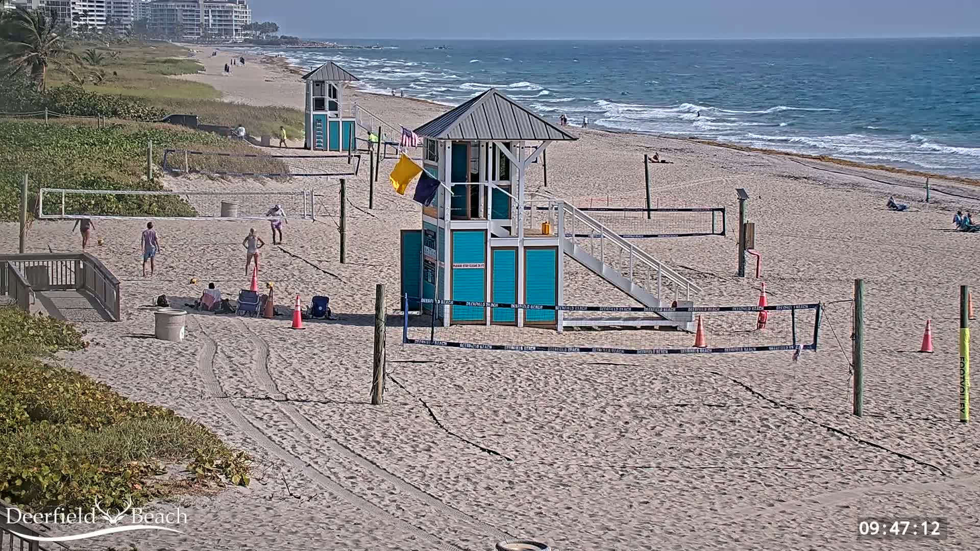 A sandy beach with volleyball nets, lifeguard stands, and a few people playing volleyball under a sunny sky with a calm ocean in the background.
