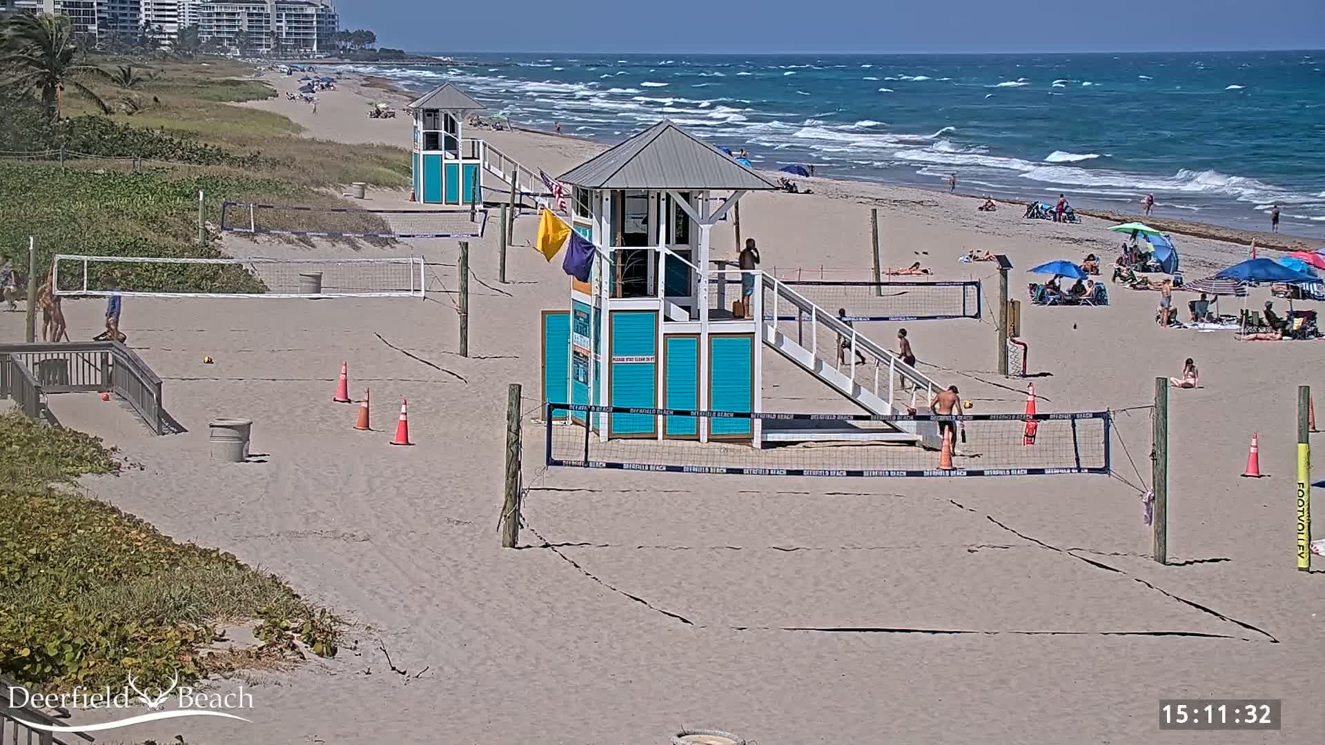 A sandy beach with volleyball courts, lifeguard stands, and scattered beachgoers under a sunny sky and a wavy ocean.