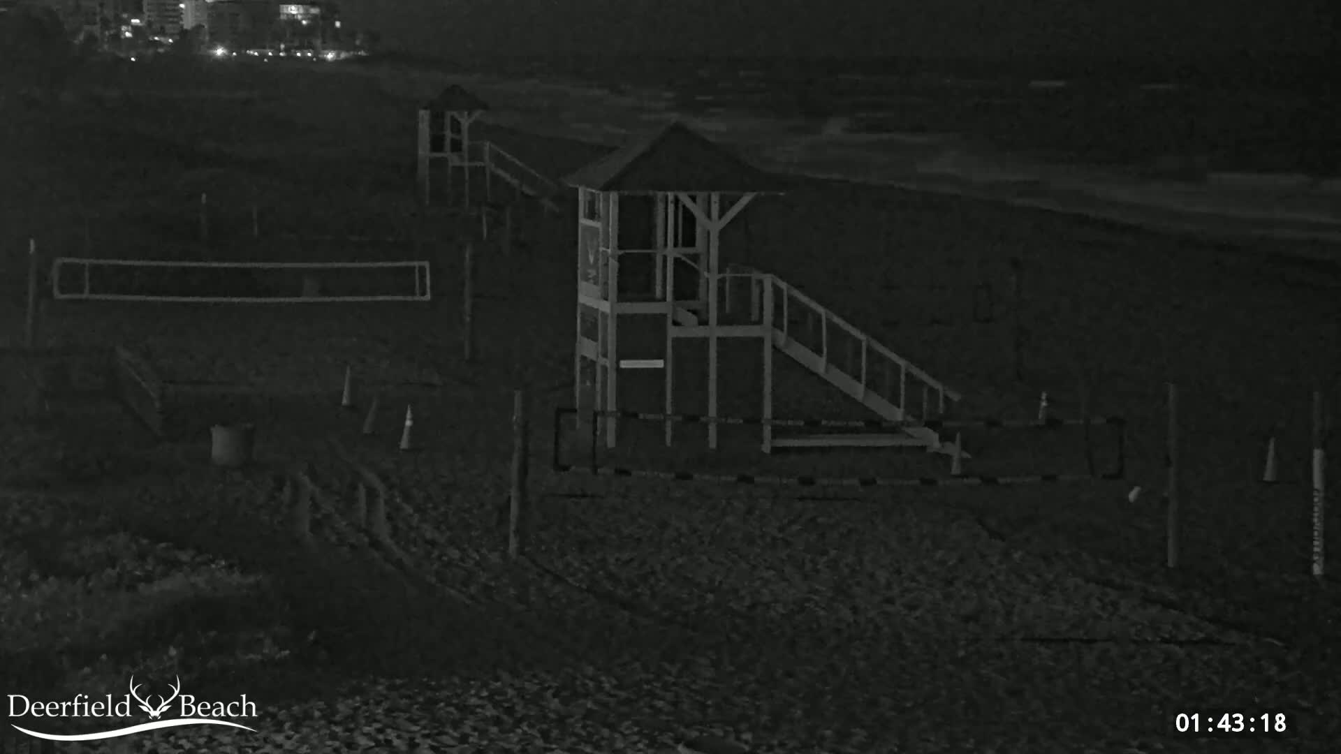 A dark, nighttime beach scene shows a lifeguard stand, volleyball nets, and some cones on a sandy beach with a dark ocean in the background.