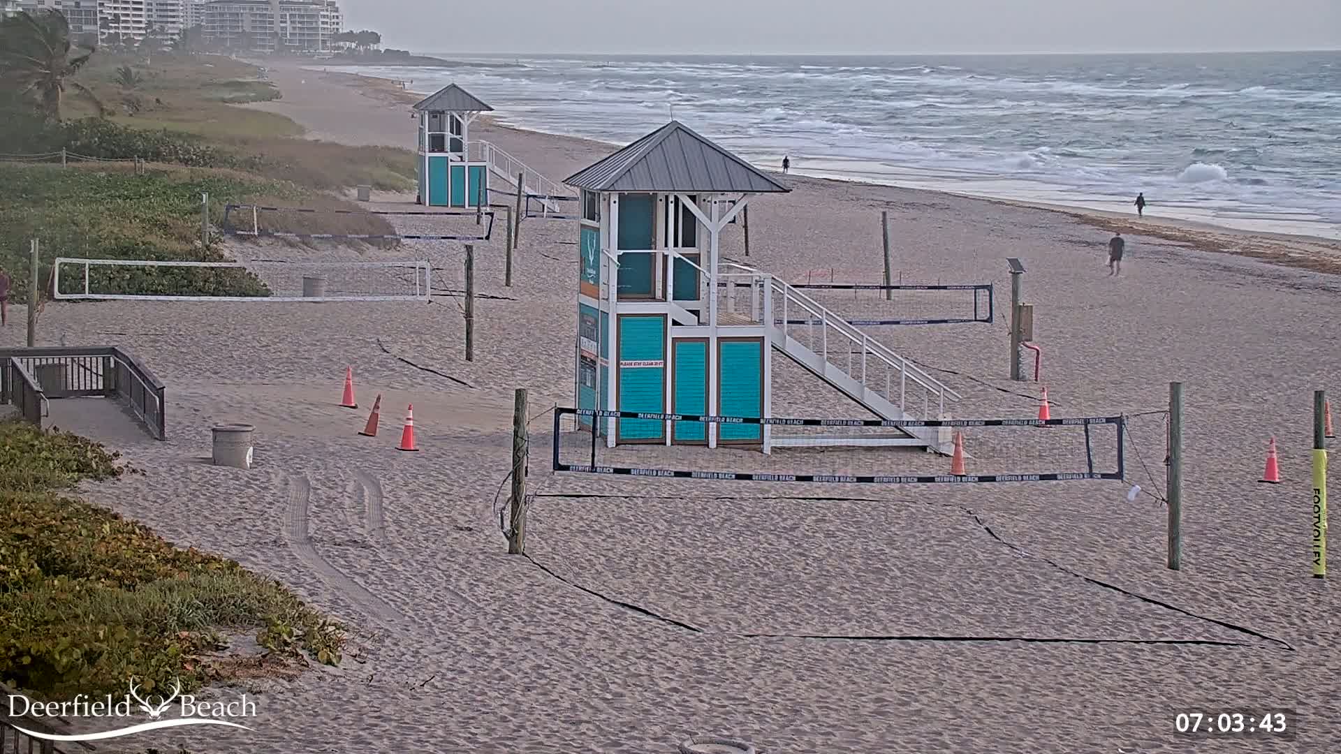 A sandy beach with several volleyball courts and lifeguard stands is shown on an overcast day with a wavy ocean in the background.