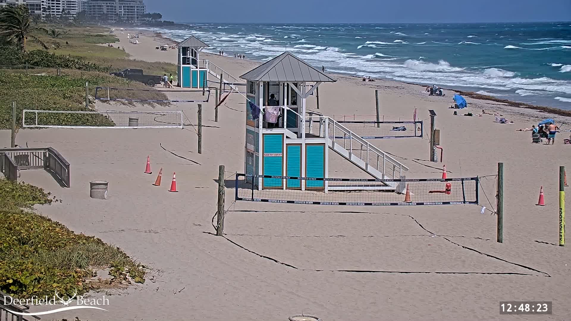A sandy beach with volleyball courts, lifeguard stands, and a few people under a partly cloudy sky and a windy ocean with waves.