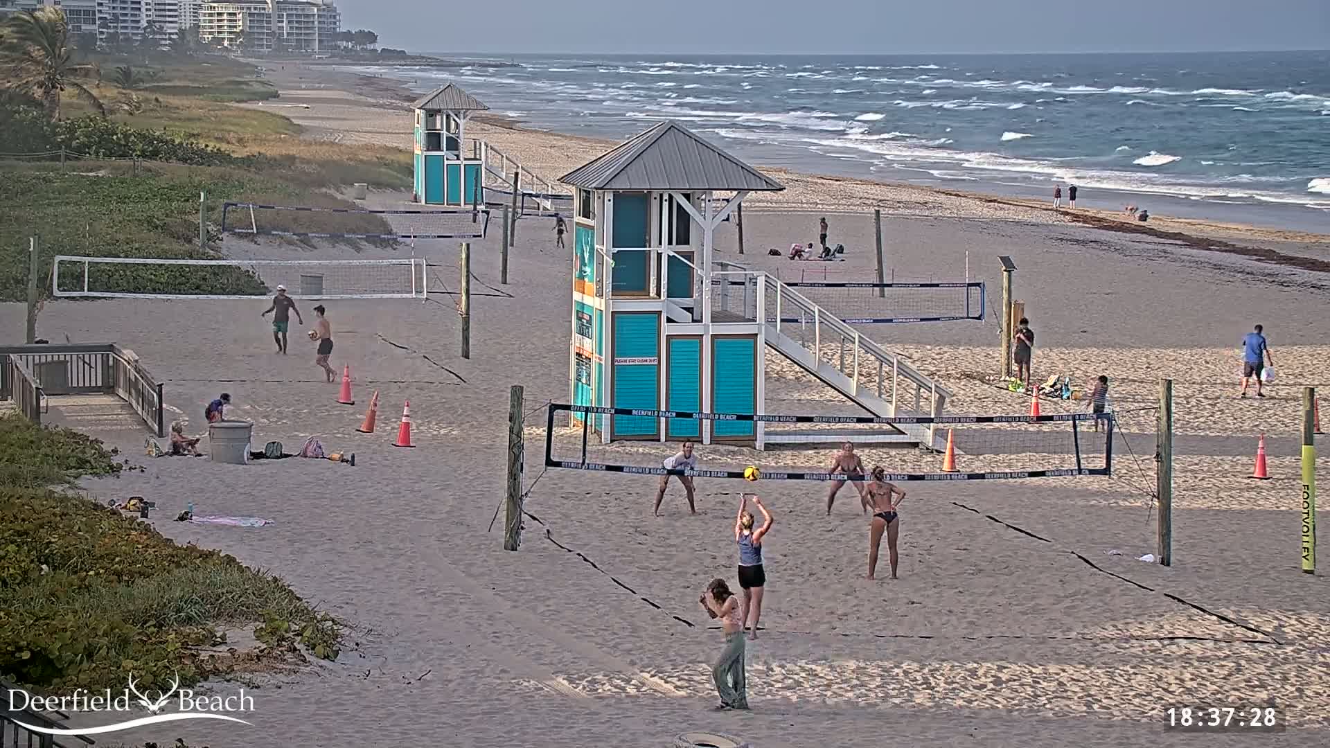 A sandy beach with several volleyball courts, lifeguard stands, and people playing volleyball and relaxing under a somewhat cloudy sky and wavy ocean.