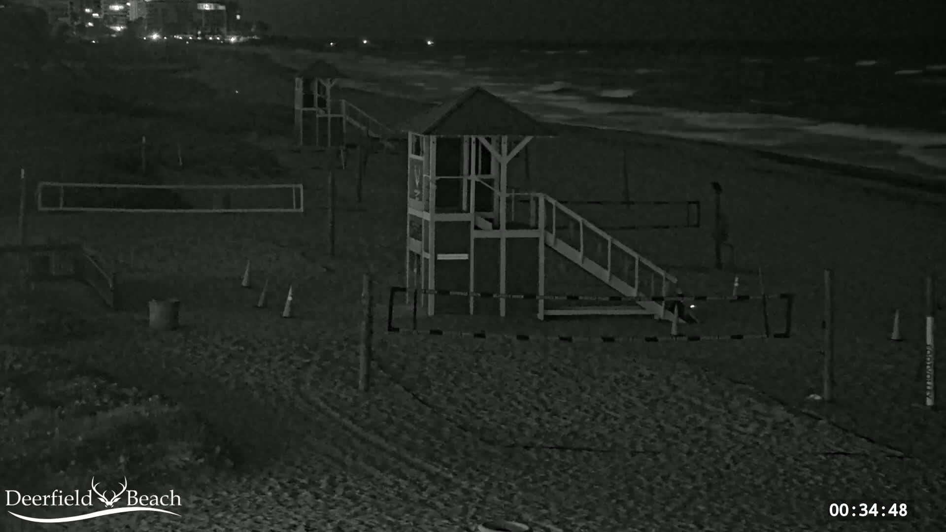 A nighttime beach scene shows a lifeguard stand, volleyball nets, and a slide, with dark, wavy water visible in the background.