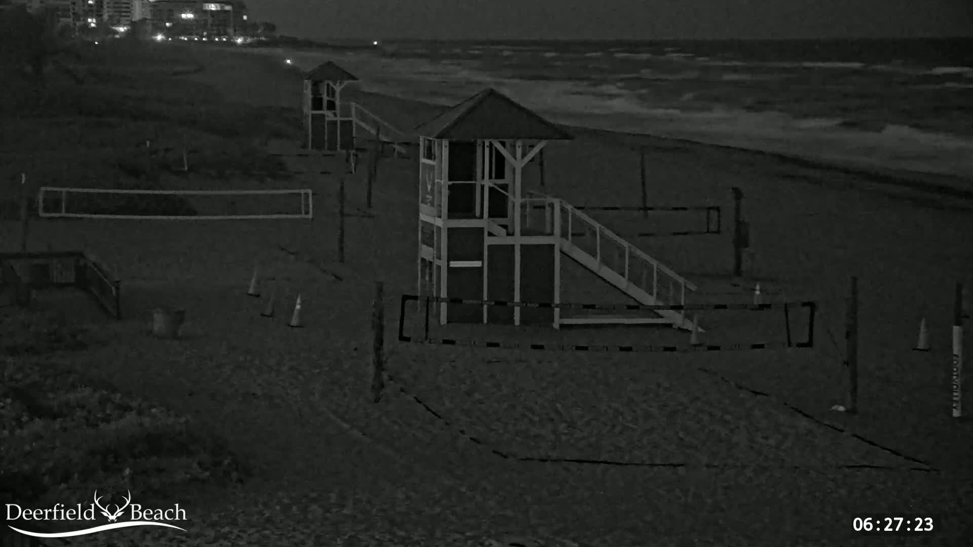 A dark, sandy beach at night shows several lifeguard stands and volleyball nets, with choppy waves visible in the background under a dark sky.