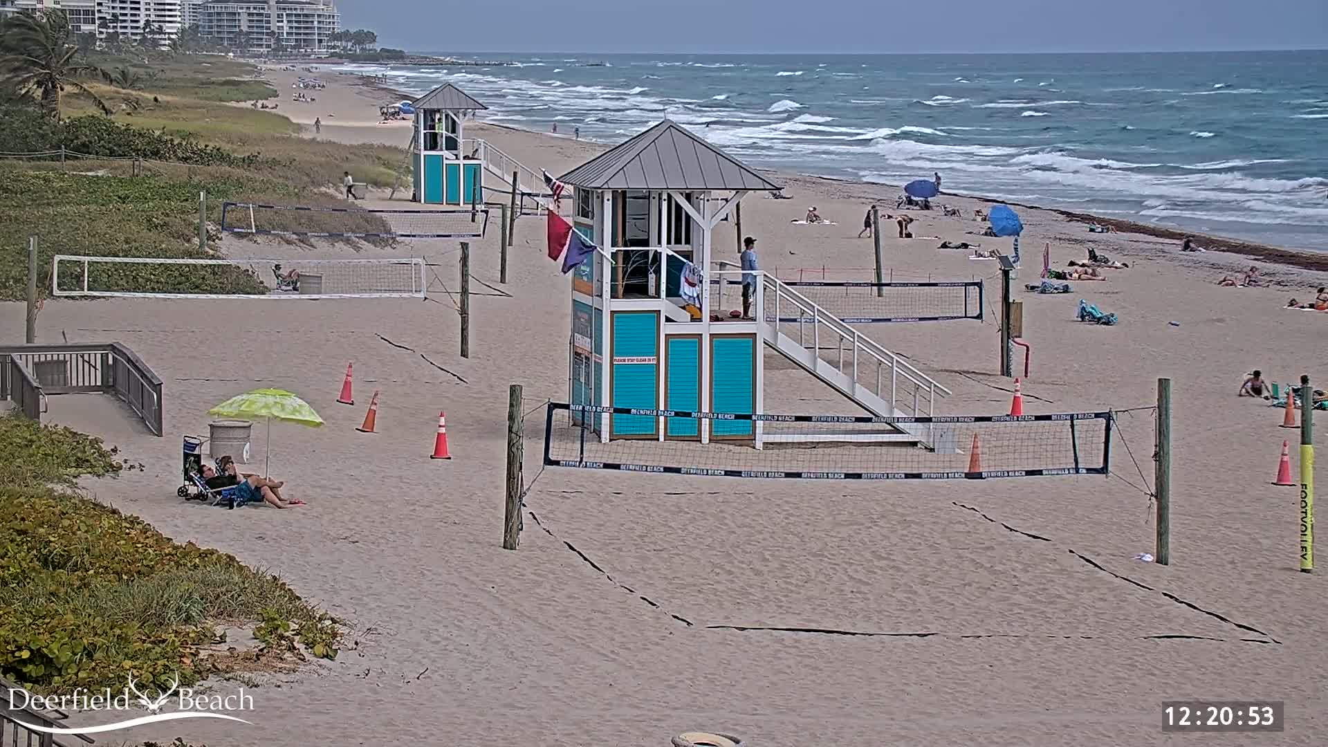 A sandy beach with lifeguard stands, volleyball courts, and a few people enjoying a partly sunny day with moderately wavy ocean waters.