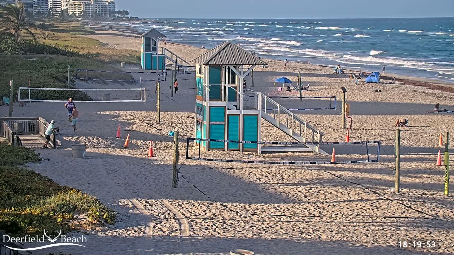 A sandy beach with volleyball courts, lifeguard stands, and a few people; the ocean has moderate waves.