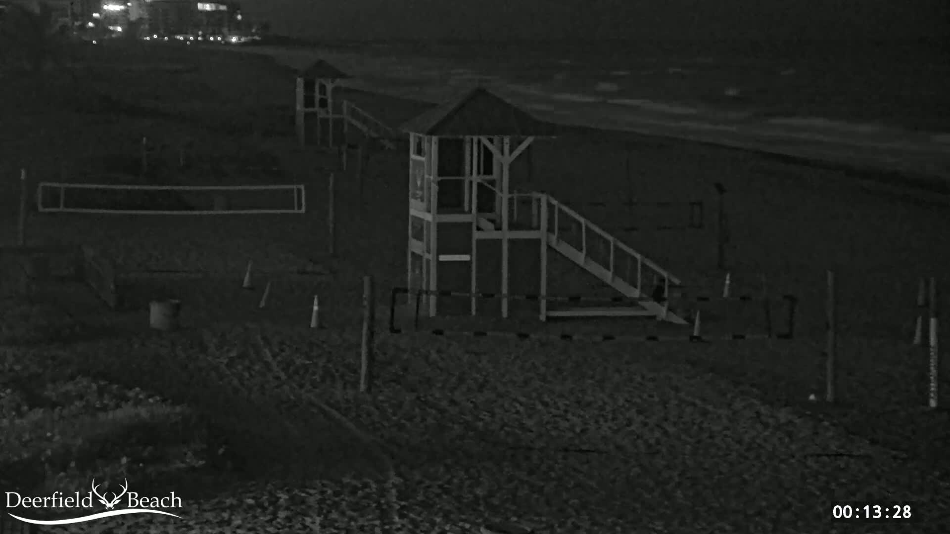 A nighttime, black-and-white image shows a beach with a lifeguard stand, volleyball net, and some other equipment, under dark, cloudy conditions.