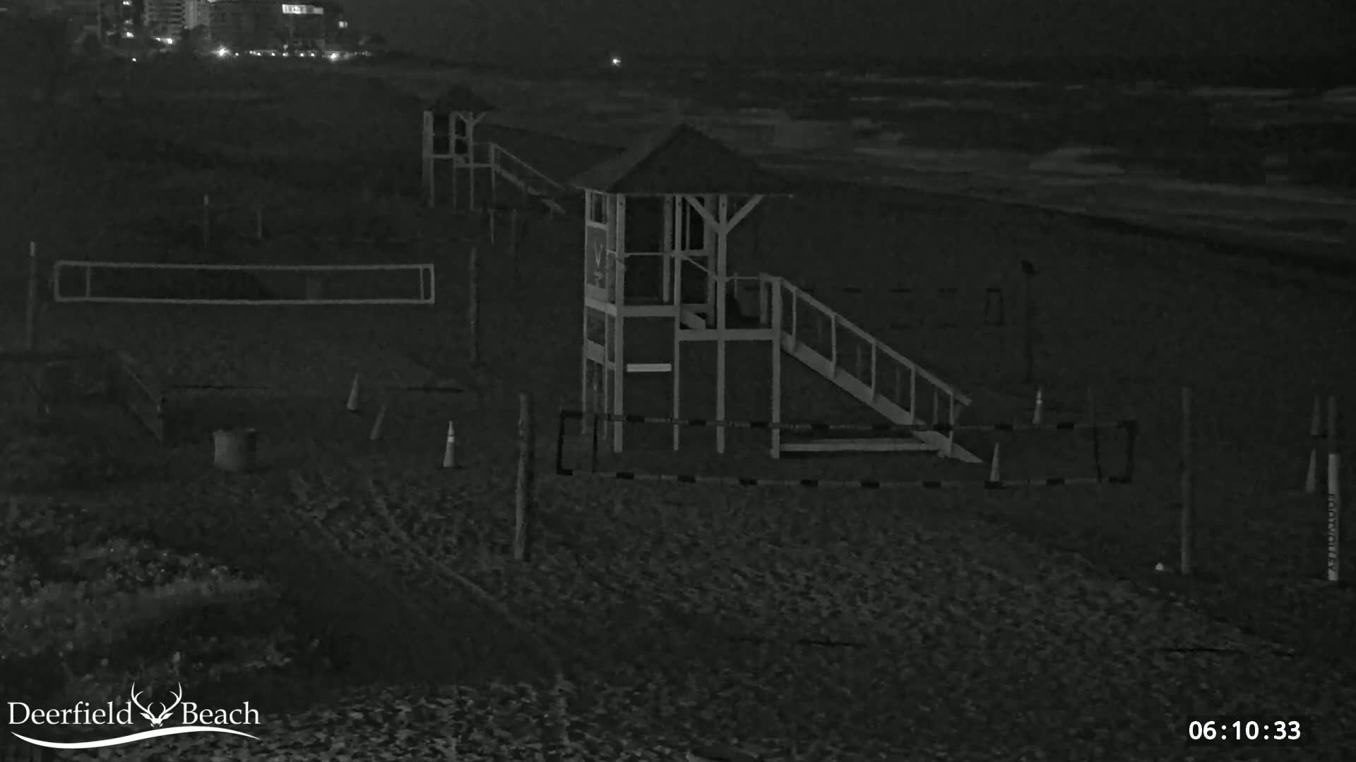 A nighttime view of a sandy beach shows a lifeguard stand, volleyball net, and other beach equipment under dark, cloudy conditions.