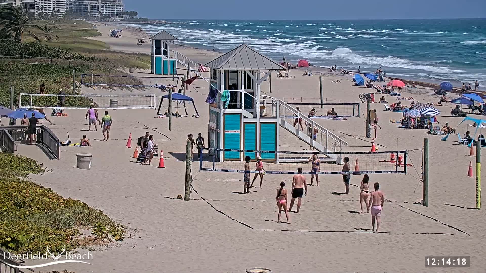 A sunny beach scene shows people playing volleyball on a sandy court near the ocean, with lifeguard stands and numerous beachgoers under umbrellas.