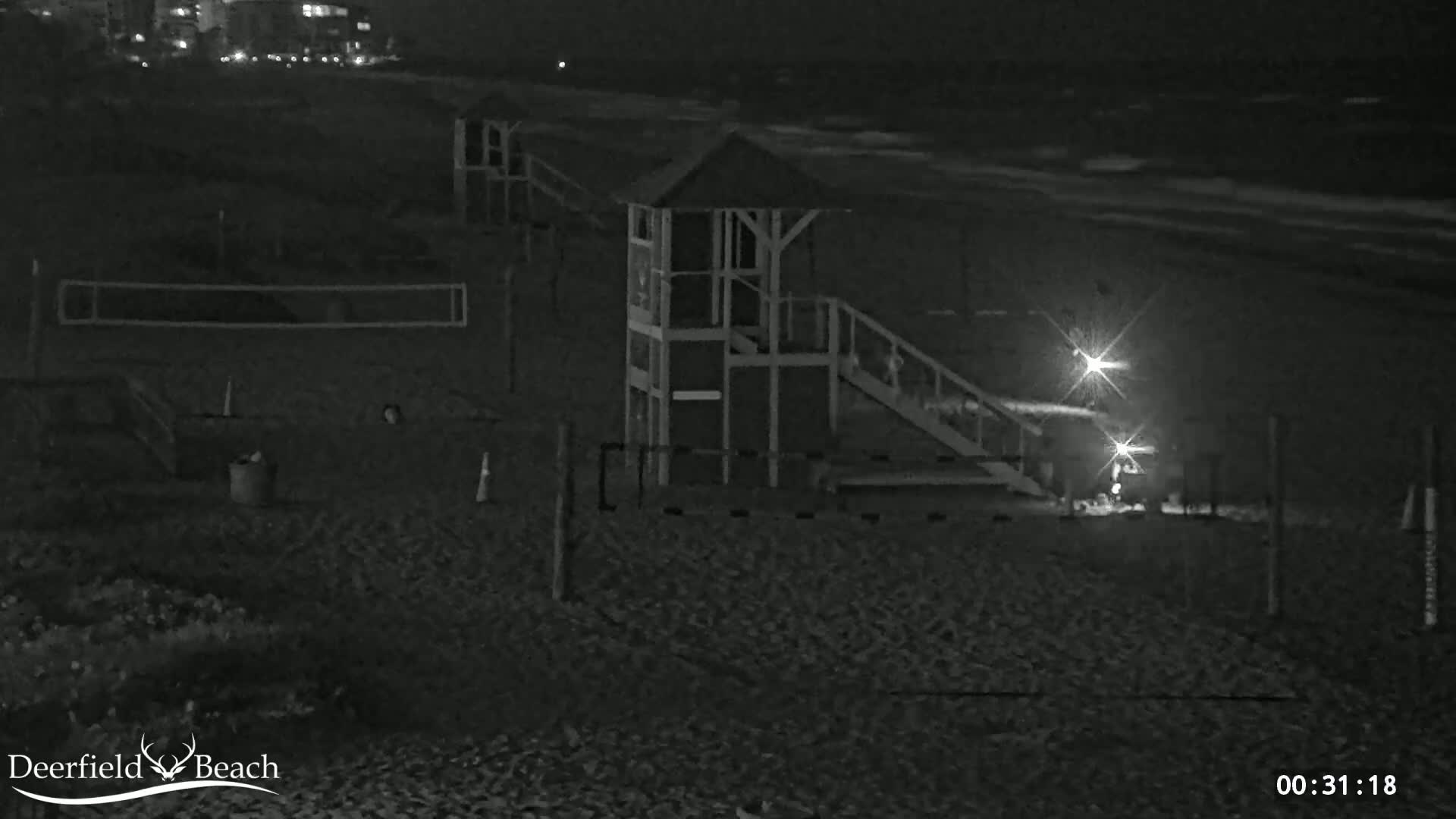 A nighttime beach scene shows an elevated lifeguard stand with a ramp, a volleyball net, and two bright lights near the ocean under dark, cloudy skies.