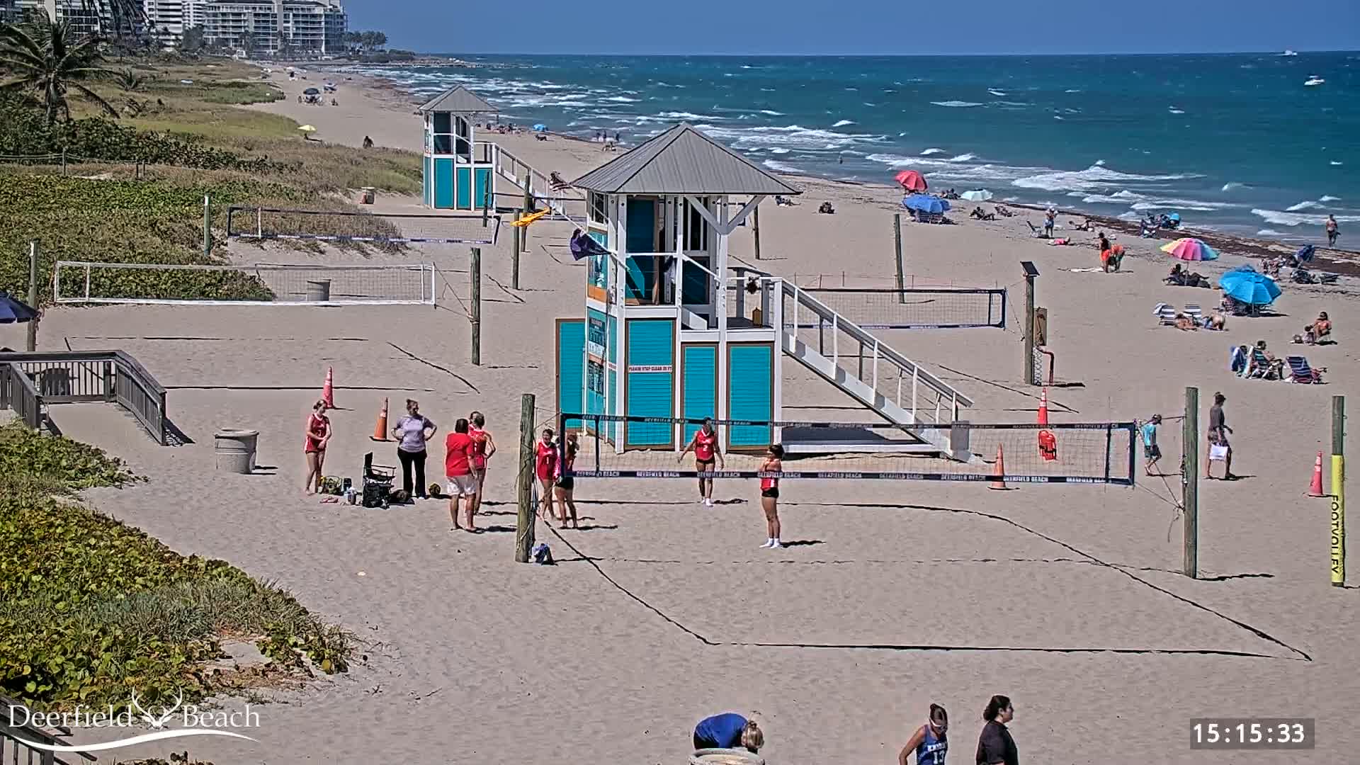 A beach volleyball game is in progress on a sandy beach under sunny skies with a windy ocean in the background.