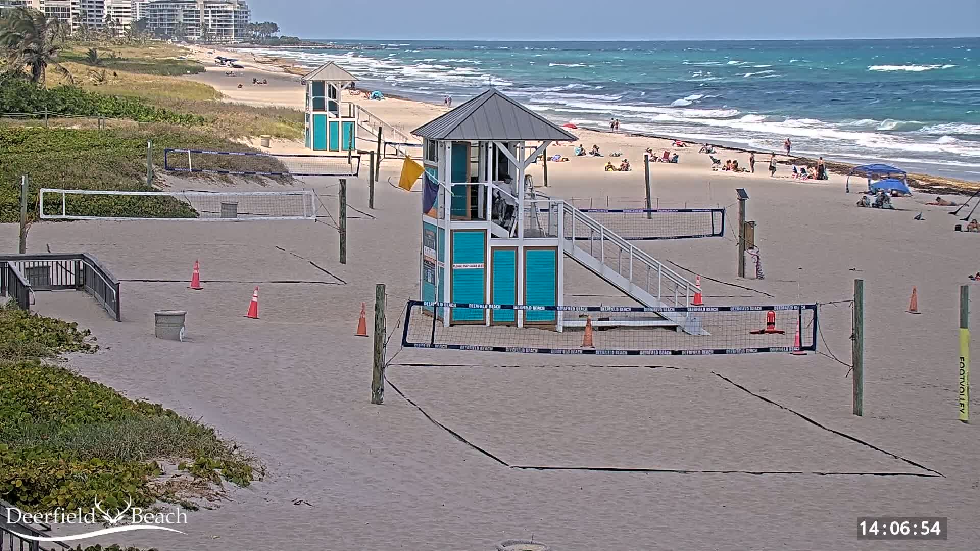 A sandy beach with volleyball courts, lifeguard stands, and a few people scattered on the shore under a partly sunny sky with a wavy ocean.
