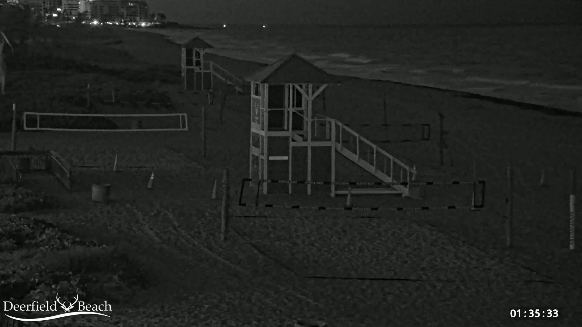 A nighttime, grayscale image shows a sandy beach with lifeguard stands, volleyball nets, and a calm ocean in the background.