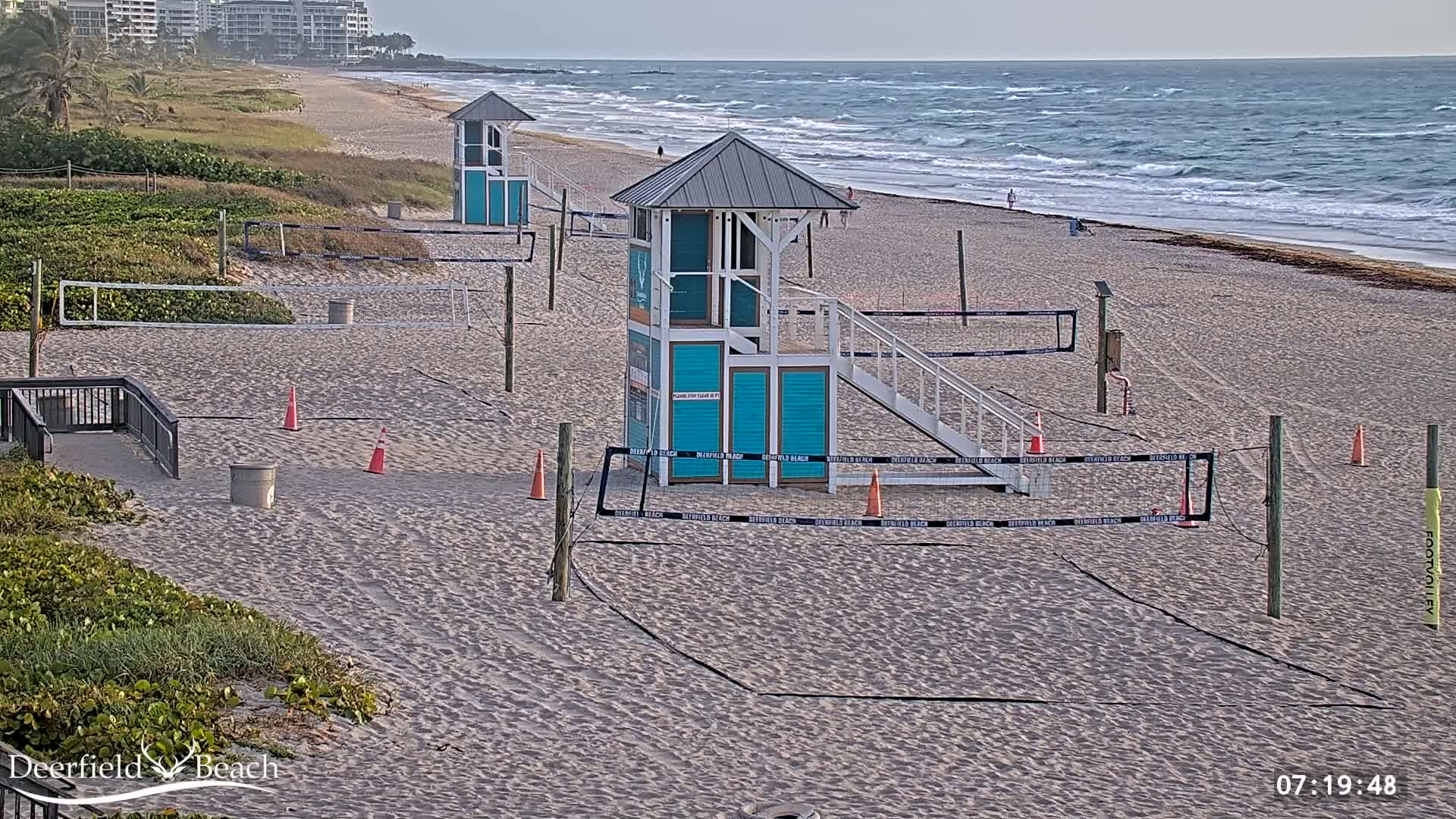 A sandy beach with several volleyball courts, lifeguard stands, and a calm ocean with small waves under a mostly clear sky.