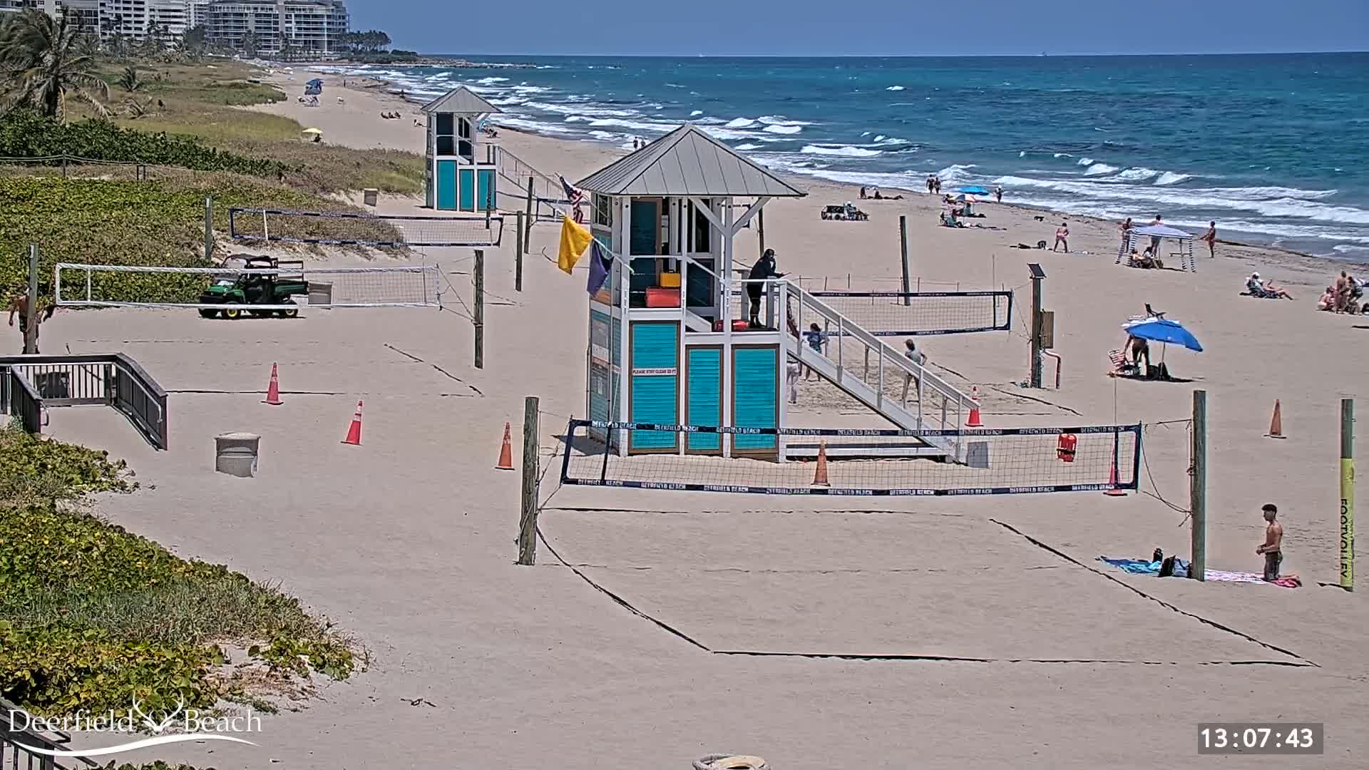 A sandy beach with volleyball courts, lifeguard stands, and a few people enjoying a sunny day with a windy ocean in the background.