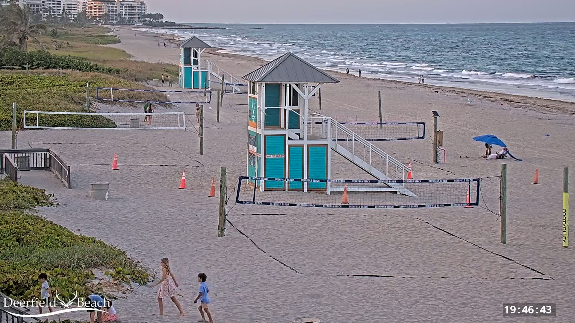 A sandy beach with several volleyball courts, lifeguard stands, and a few people, under a mostly clear sky with a calm ocean in the background.