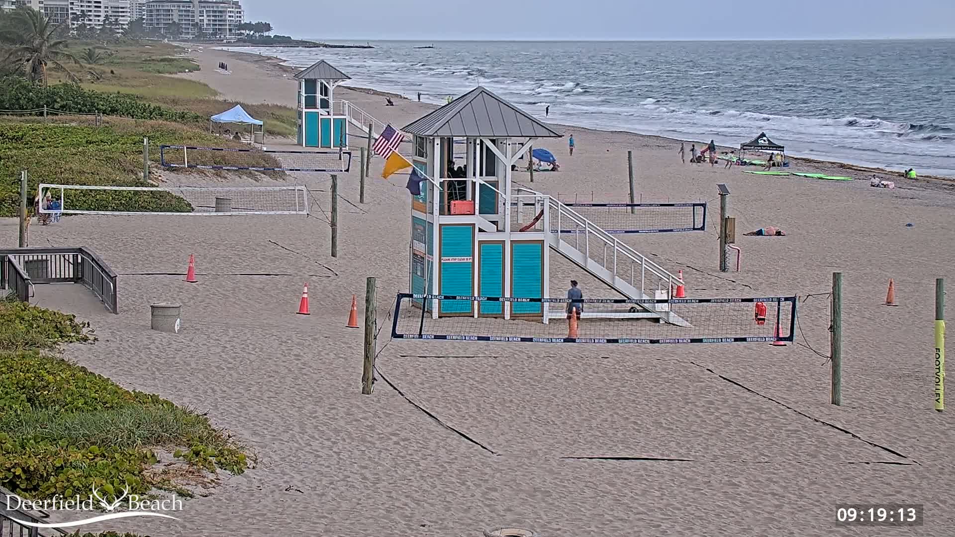 A sandy beach with several volleyball courts, lifeguard stands, and a few people under an overcast sky and moderate waves.