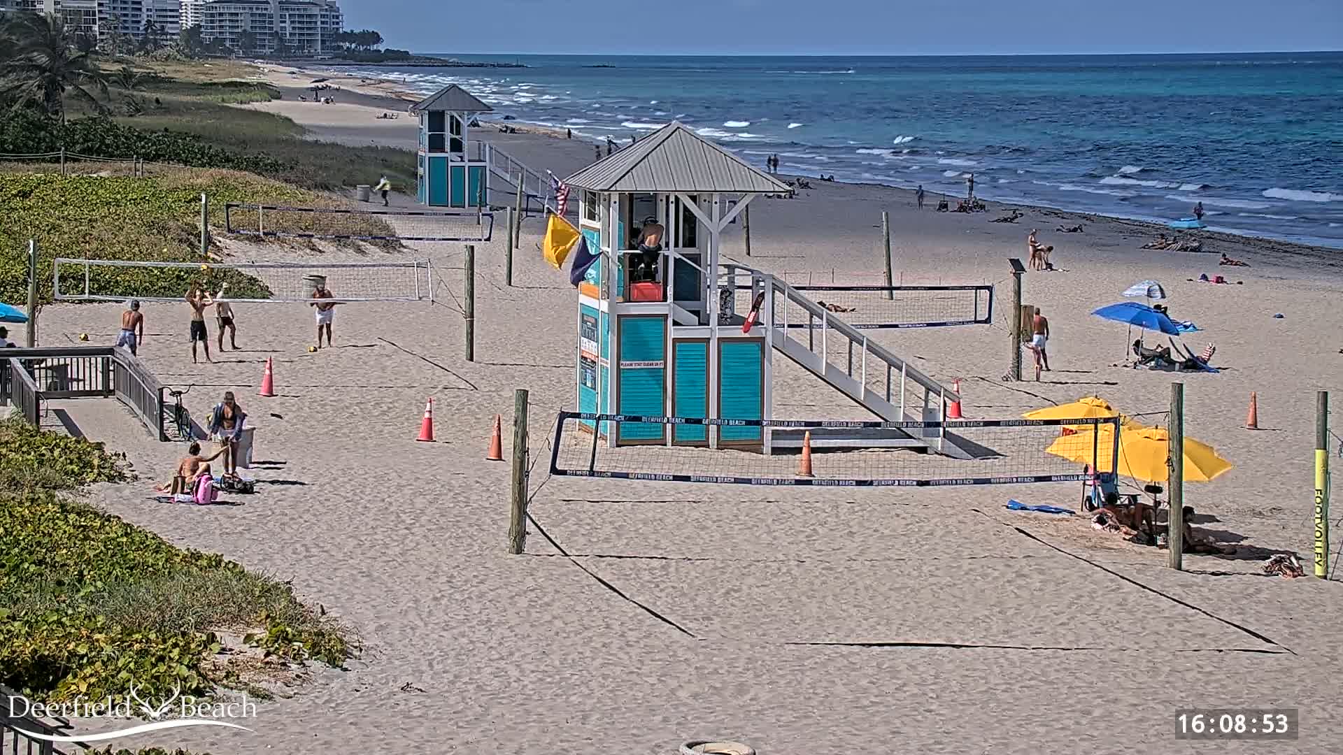 A sandy beach with volleyball courts, lifeguard stands, and several people enjoying a sunny day.
