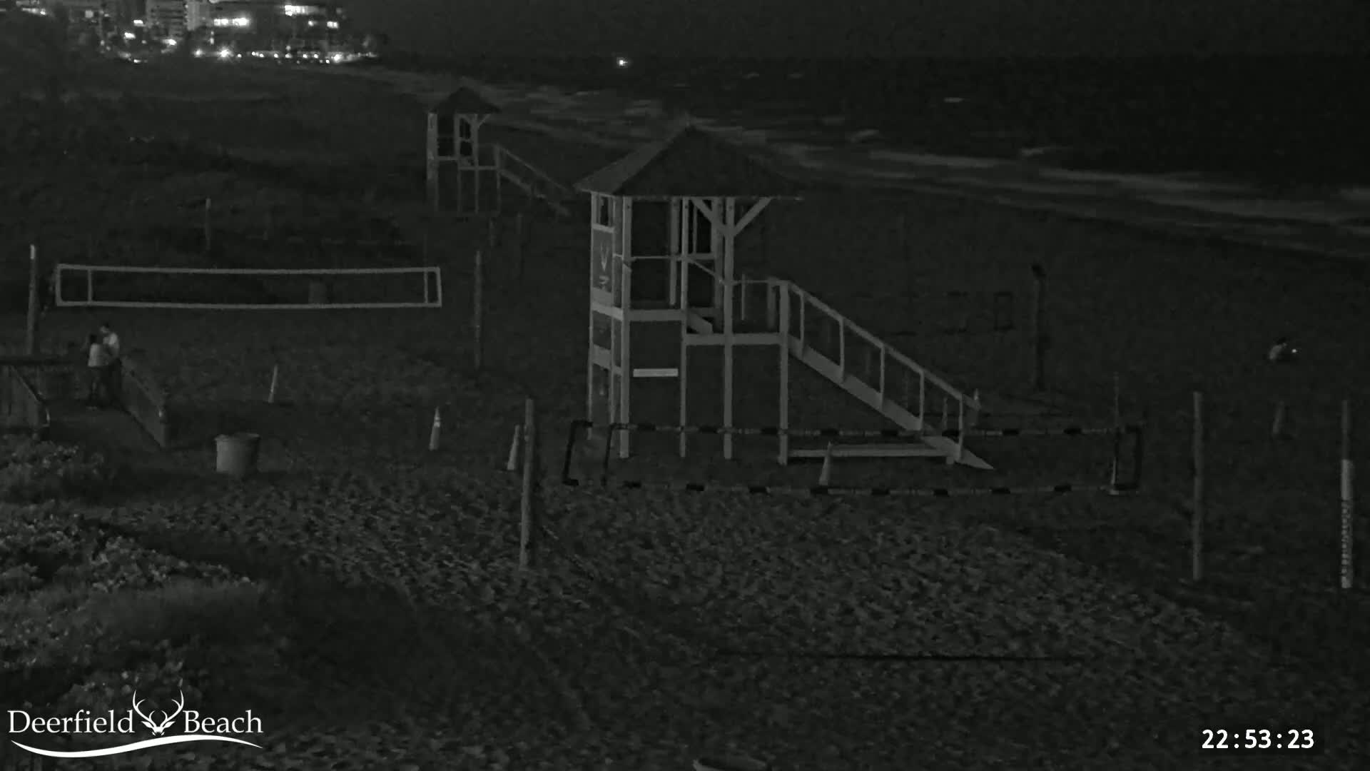 A nighttime beach scene shows a lifeguard stand, volleyball net, and two people near the net, with dark, gently lapping waves in the background.
