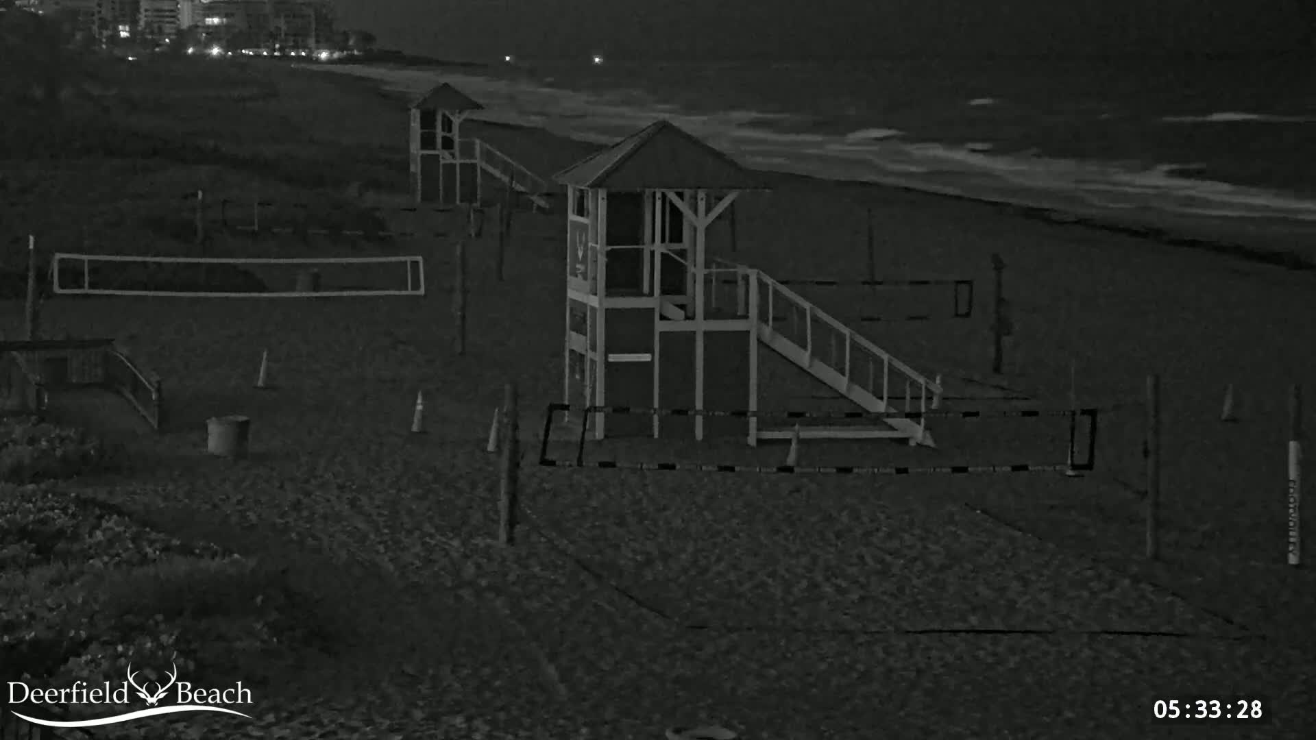 A nighttime beach scene shows a volleyball net, lifeguard stands, and gently lapping waves under a dark sky.