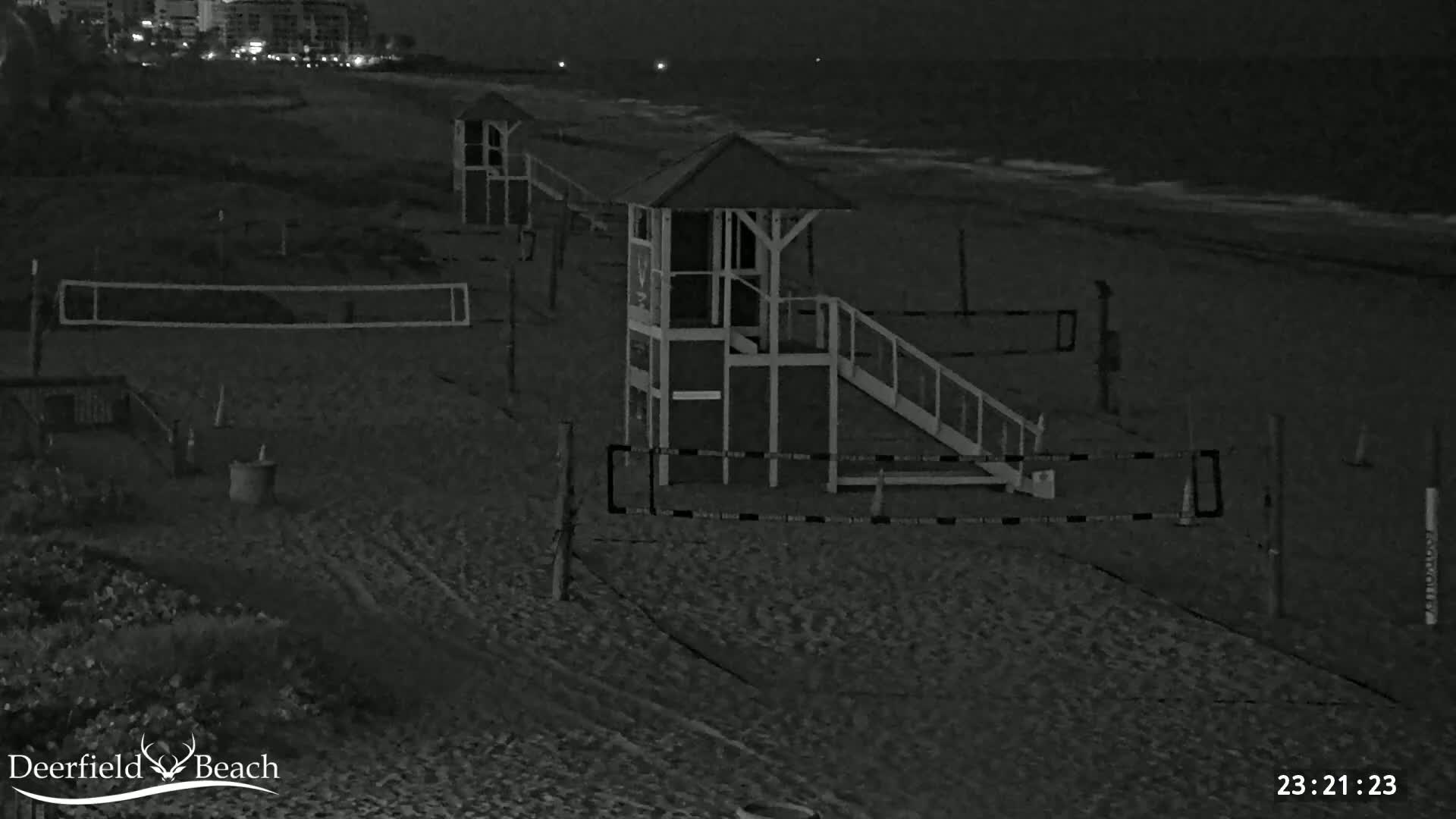 A nighttime, black-and-white view of a sandy beach with lifeguard stands, volleyball nets, and other beach equipment under dark, nighttime conditions.