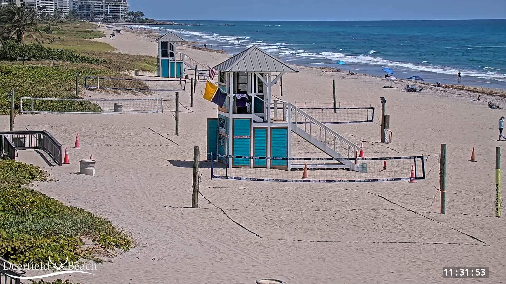 A sandy beach with volleyball courts, lifeguard stands, and a few people visible on a sunny day with light waves in the ocean.