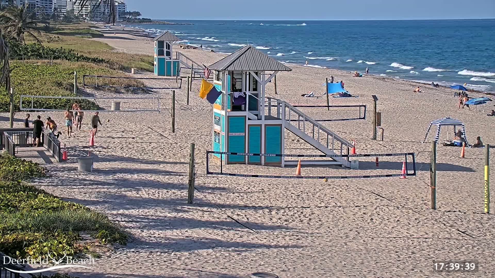 A sandy beach with volleyball courts, lifeguard stands, and several people enjoying a sunny day with calm ocean waves.