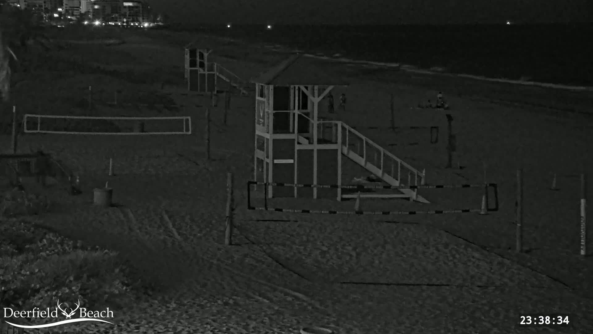 A dark, sandy beach at night shows several volleyball nets and lifeguard stands, with a few people visible near the water.
