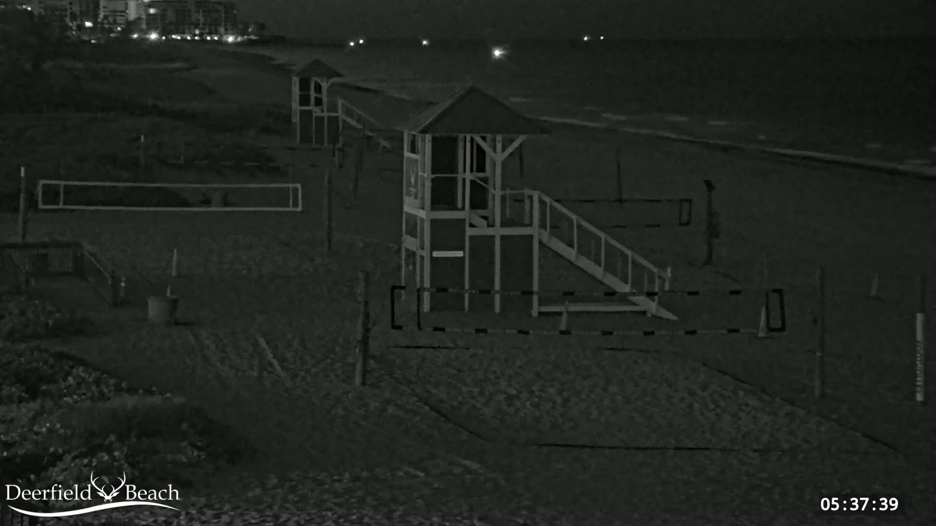 A nighttime, grayscale image shows a sandy beach with lifeguard stands, volleyball nets, and a single person standing near the water's edge under dark, cloudy skies.