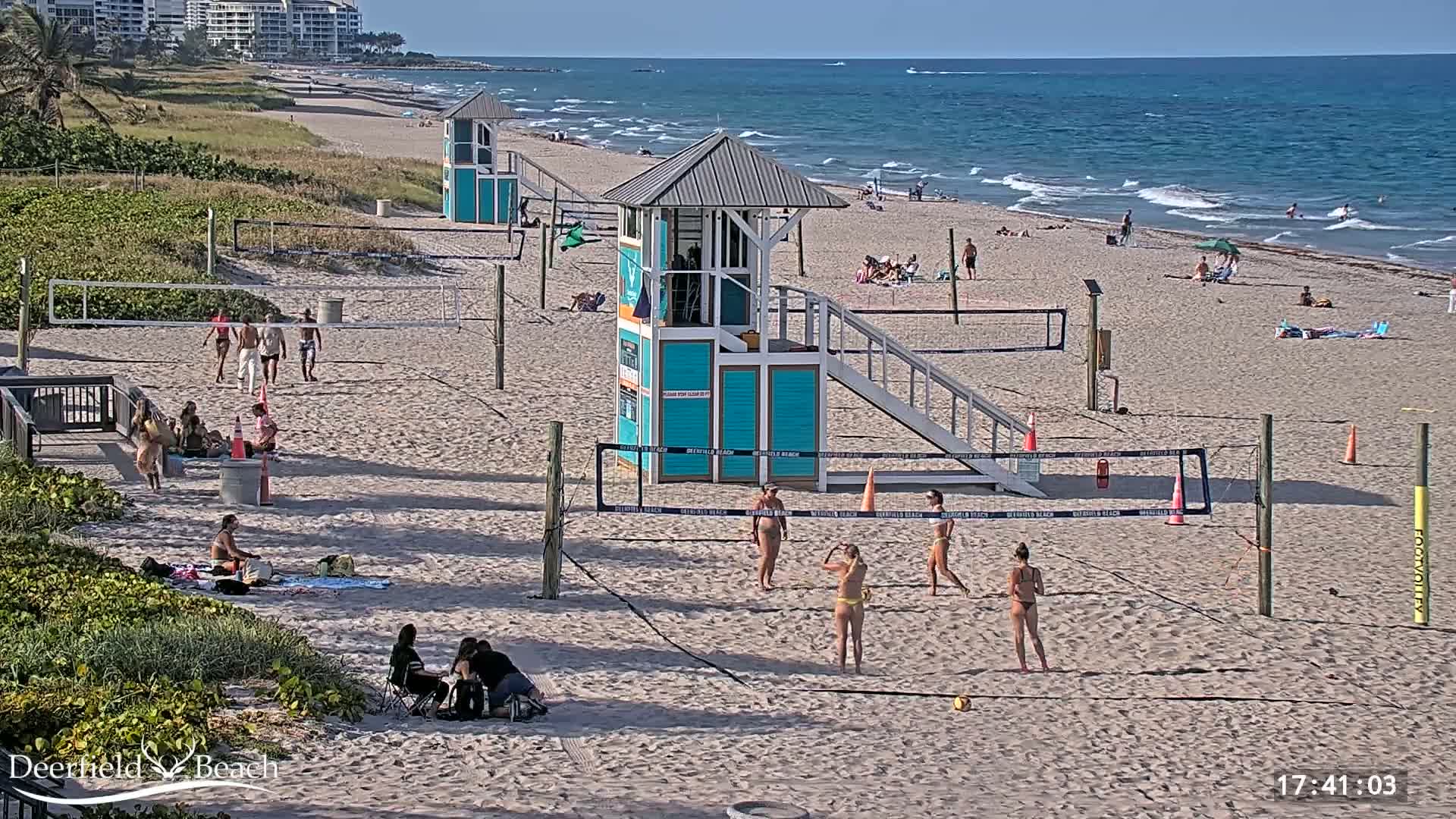 A sandy beach with several volleyball courts, lifeguard stands, and people playing volleyball and relaxing under a sunny sky with a calm ocean in the background.