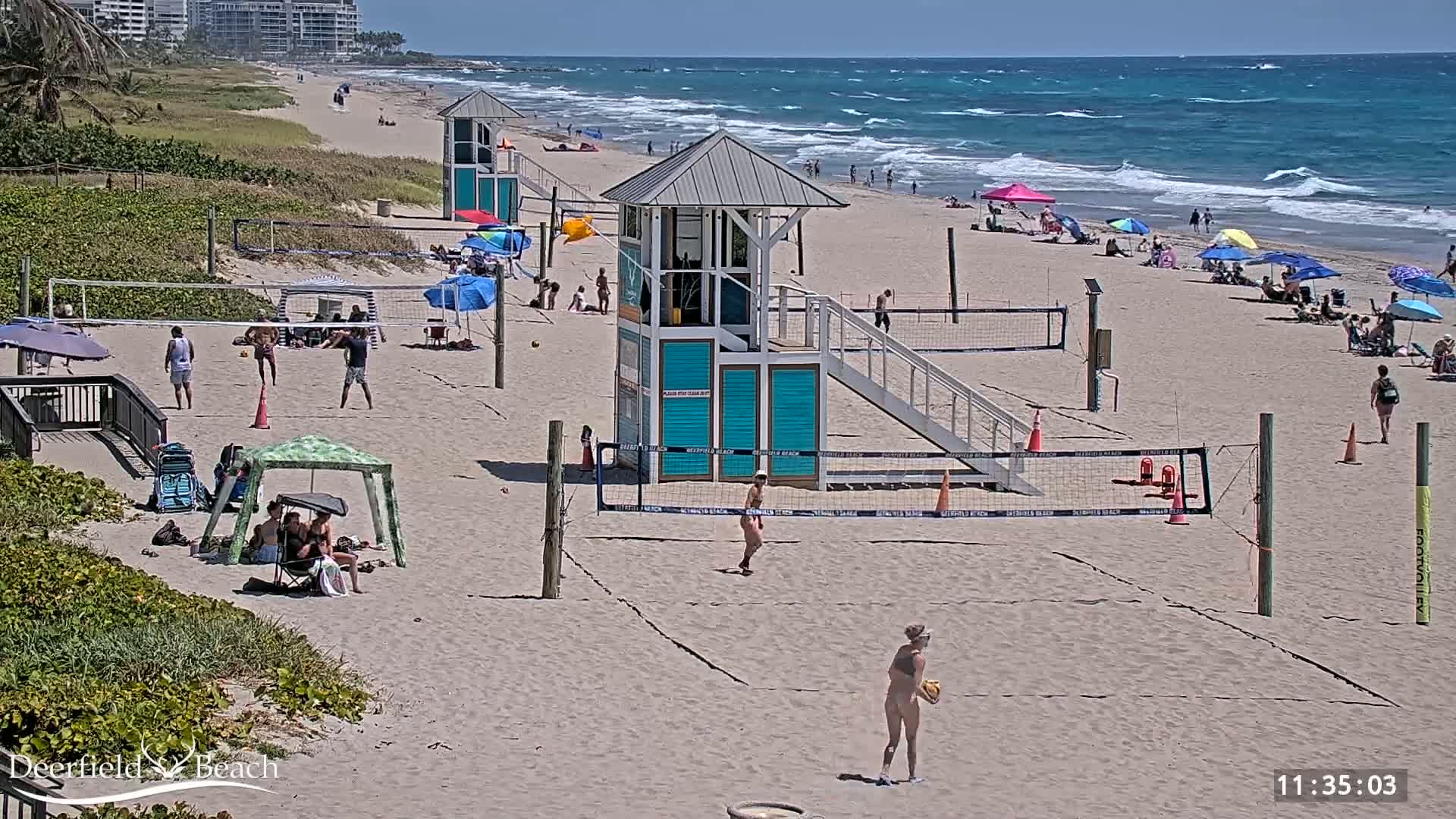 A sandy beach with volleyball courts, lifeguard stands, and beachgoers enjoying a sunny day with a windy ocean in the background.