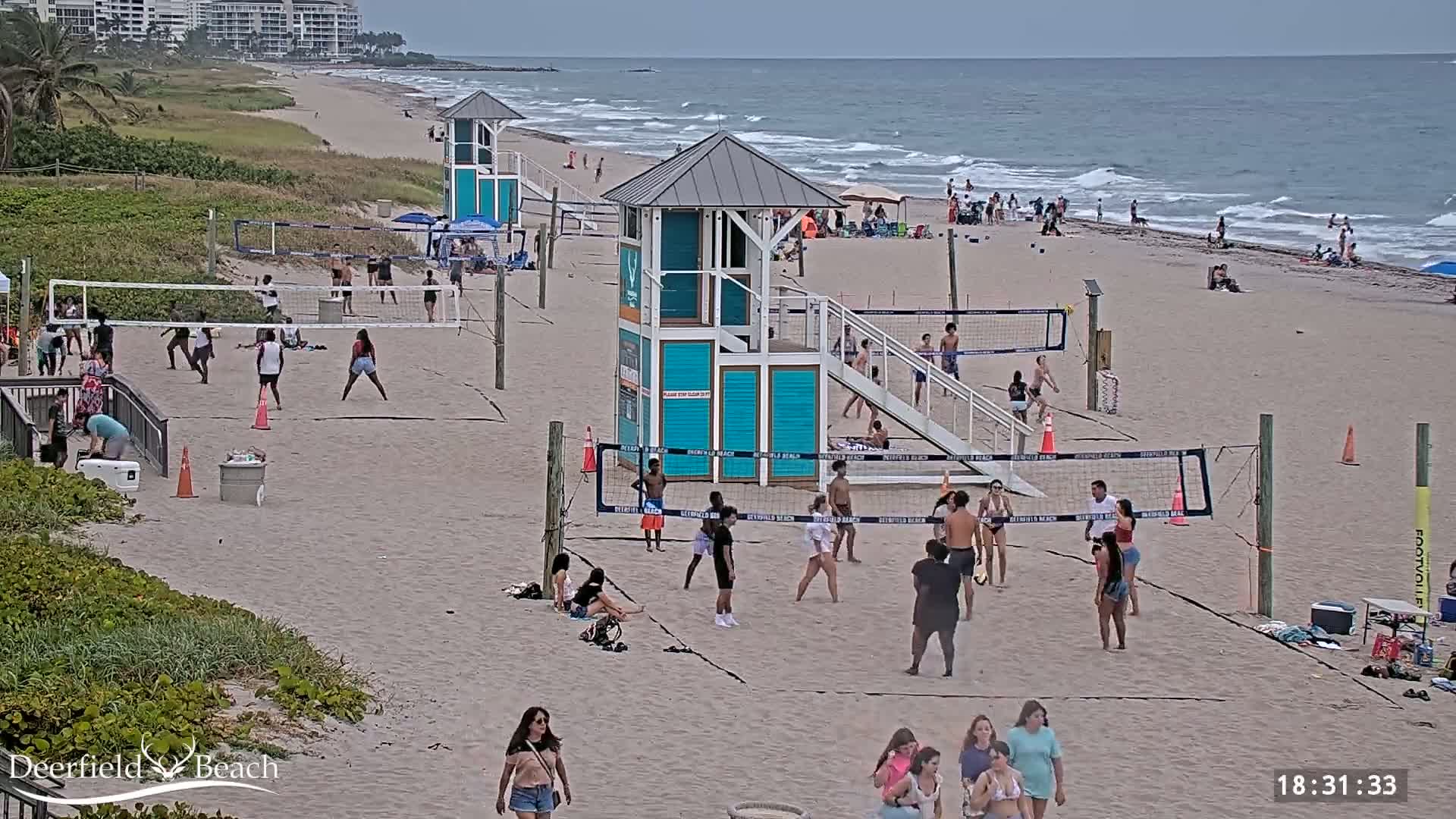 A sandy beach with several volleyball courts, numerous people playing volleyball and relaxing, and a lifeguard stand; the ocean is visible in the background on an overcast day.