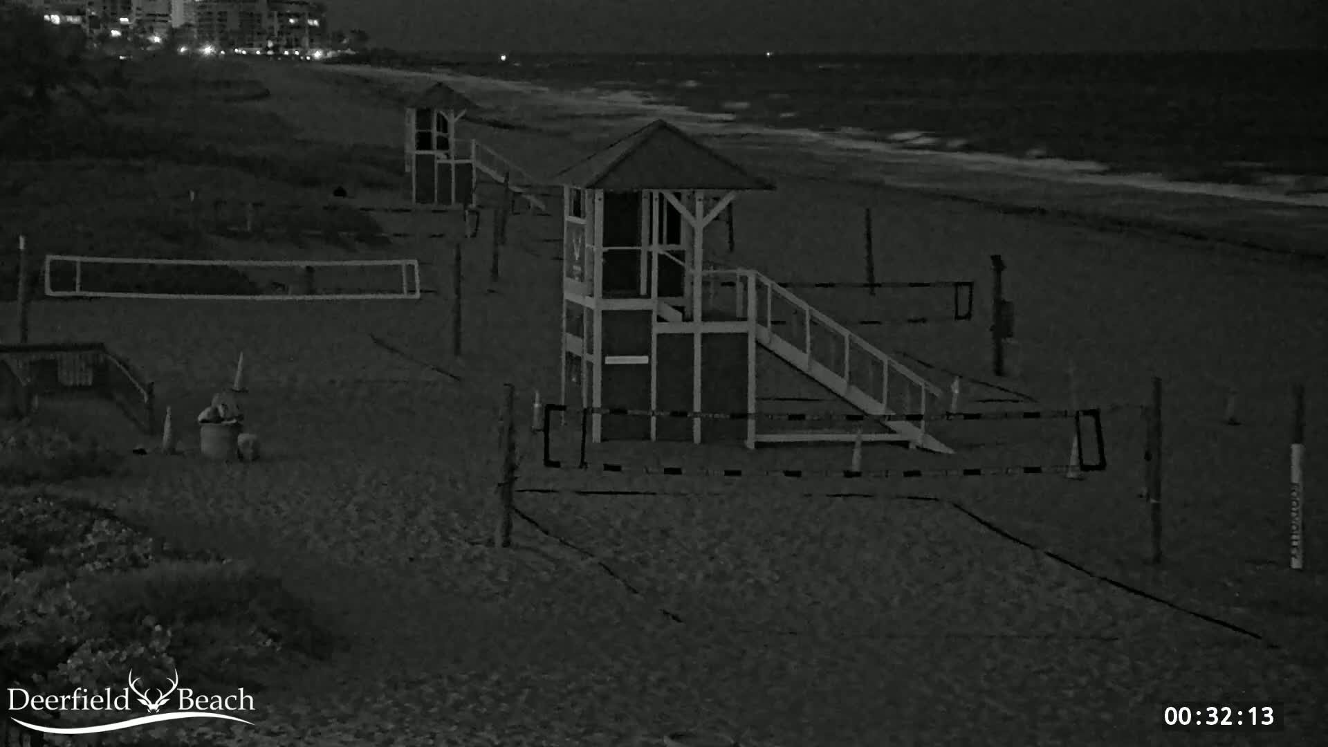 A dark, sandy beach at night shows several lifeguard stands, volleyball nets, and a single person sitting near some beach equipment under a dark sky with a slightly wavy ocean in the background.