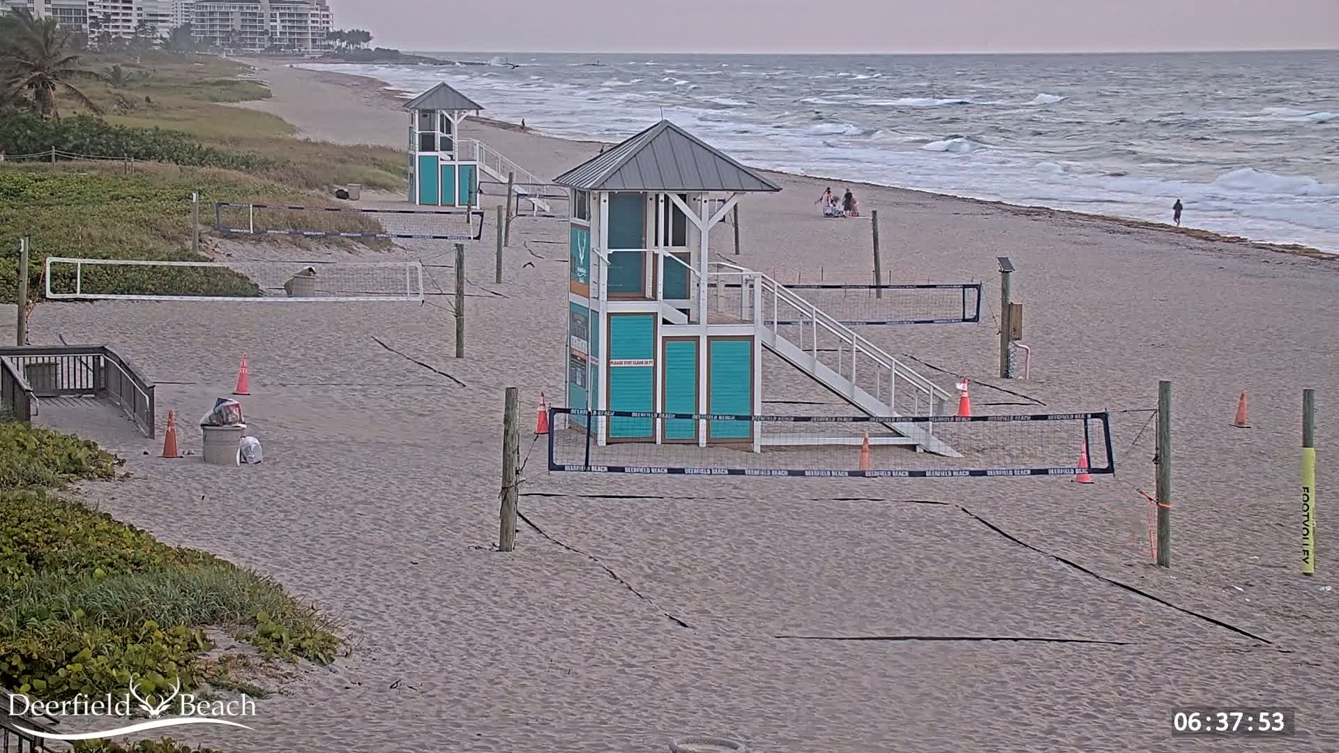 A sandy beach with several volleyball courts and lifeguard stands, under an overcast sky and with a wavy ocean in the background.