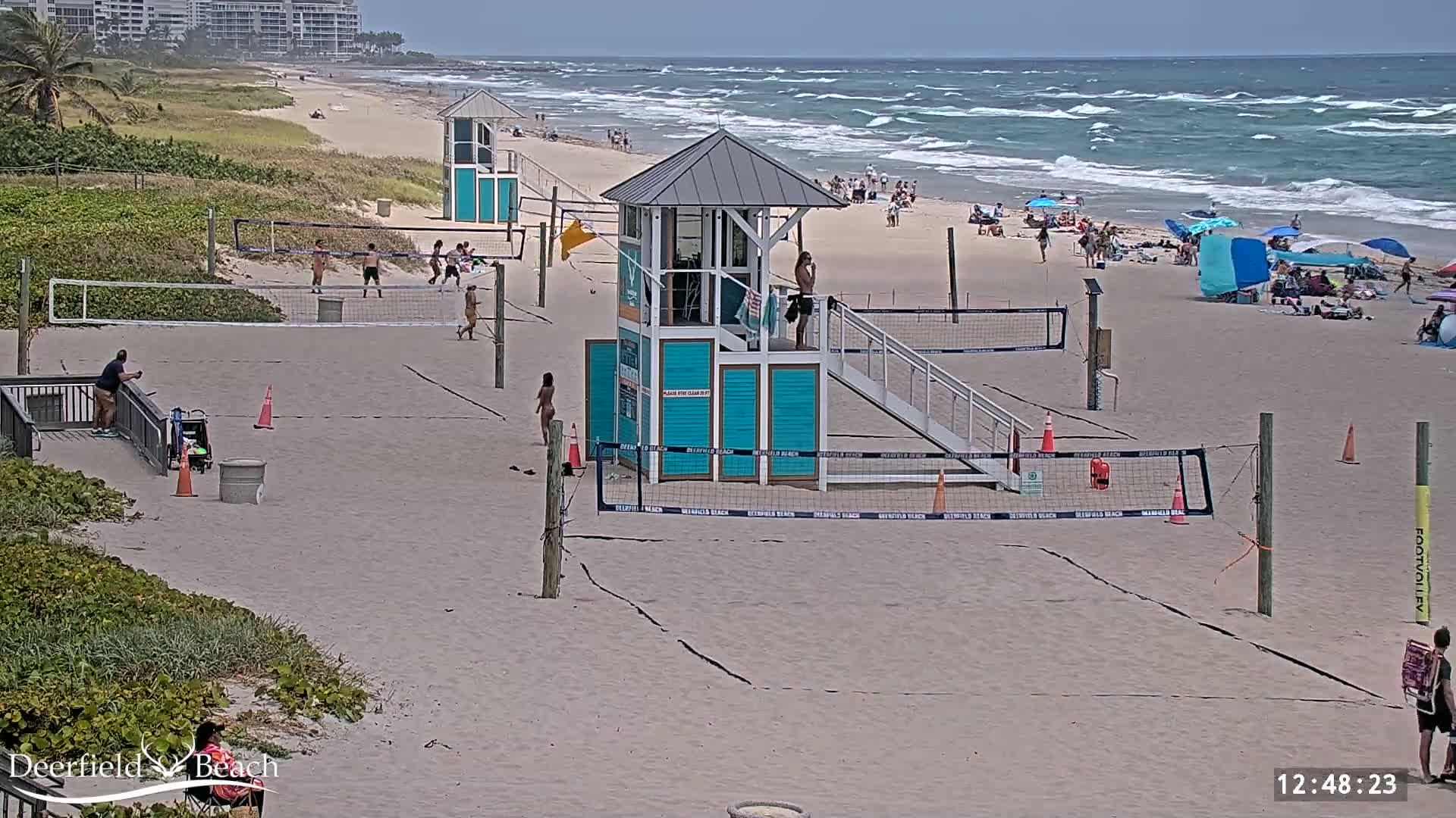 A sandy beach with volleyball courts, lifeguard stands, and people enjoying the beach on a partly cloudy day with moderate waves.