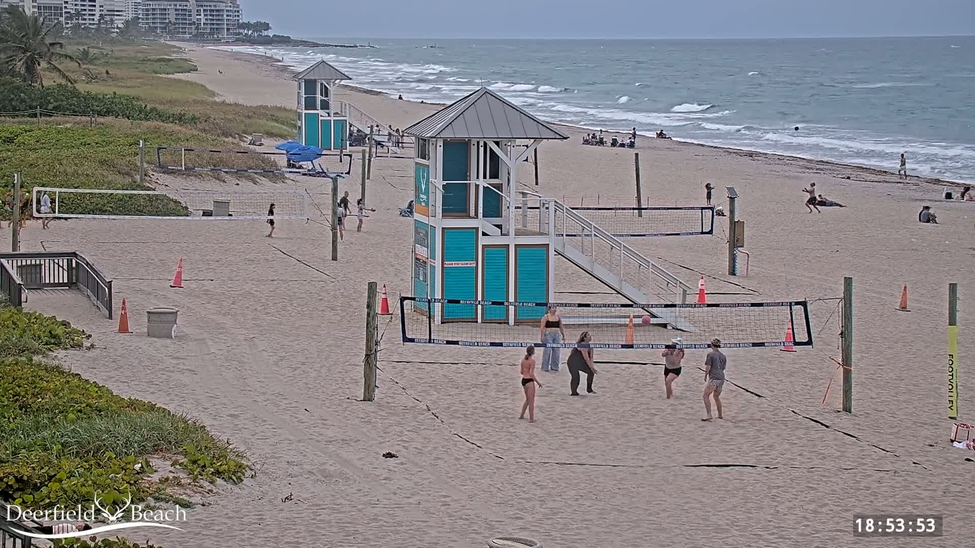 A sandy beach with several volleyball courts, lifeguard stands, and people playing volleyball under partly cloudy skies and a somewhat choppy ocean in the background.