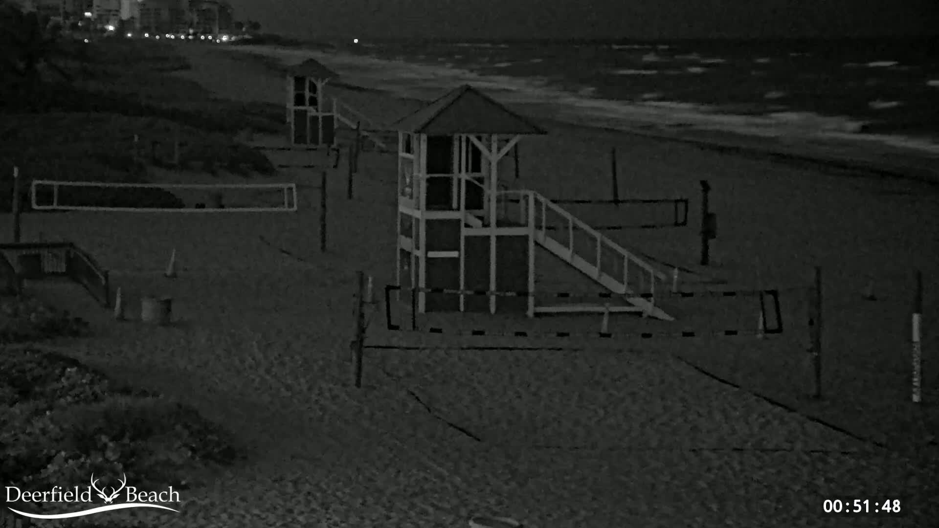 A dark sandy beach at night shows three lifeguard stands and volleyball nets, with a slightly wavy ocean in the background under a dark sky.