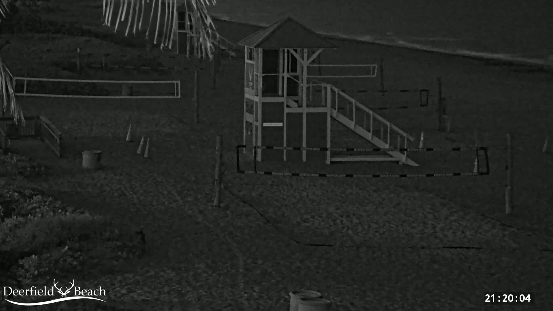 A nighttime, low-light image shows a beach with a lifeguard stand, volleyball net, and a slide, under dark, cloudy conditions.