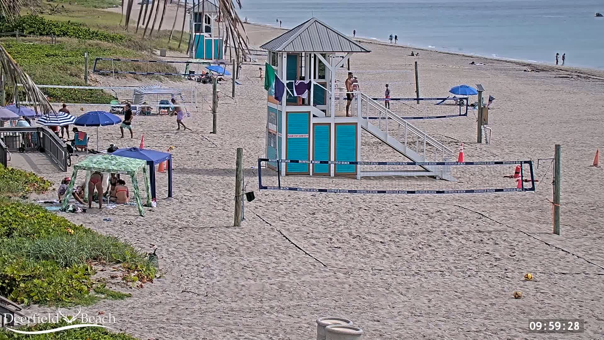 A sandy beach with volleyball courts, lifeguard stands, and several people enjoying the sunny day.