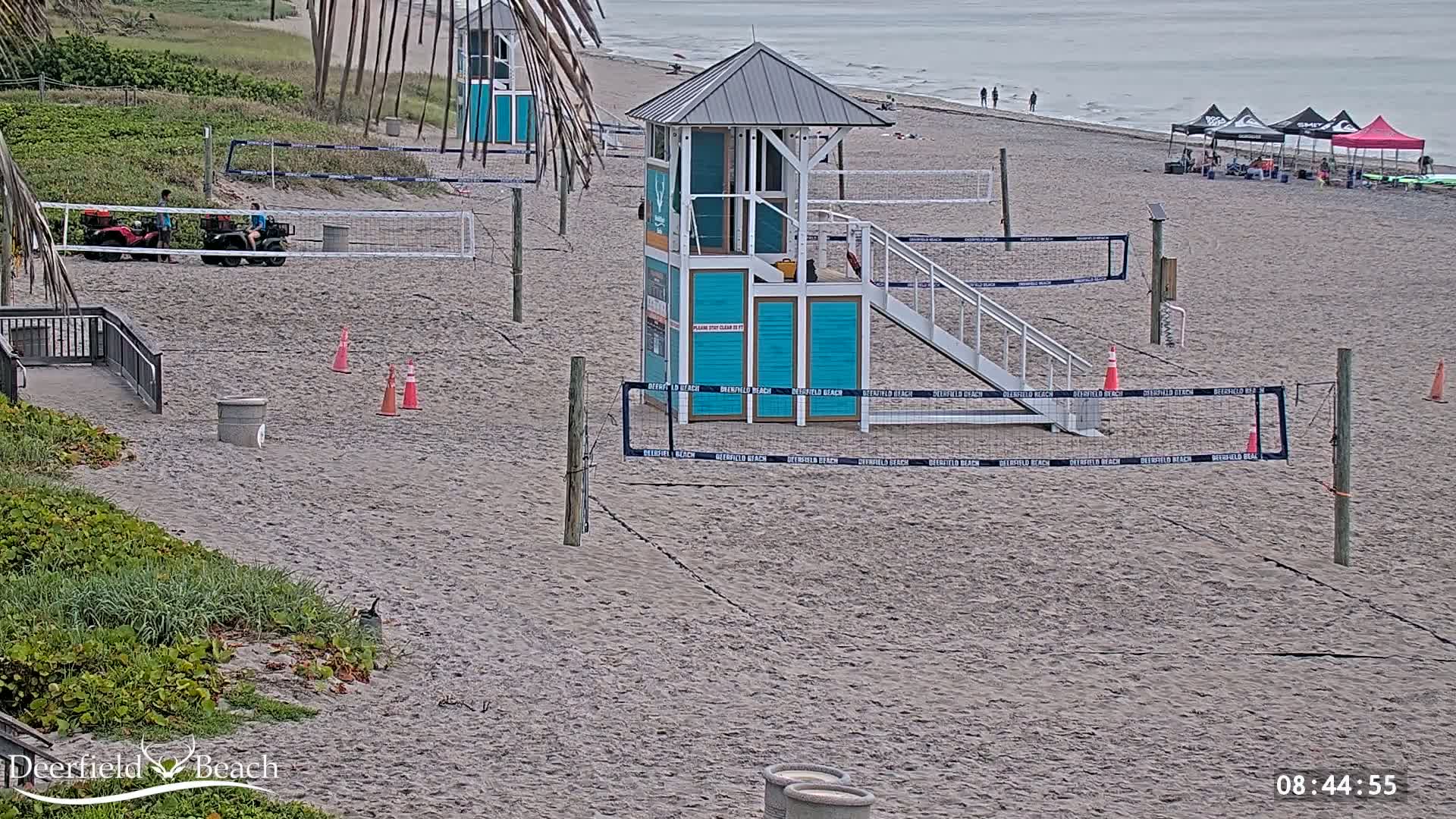 A sandy beach with volleyball courts, a lifeguard stand, and two utility vehicles is shown on an overcast day.
