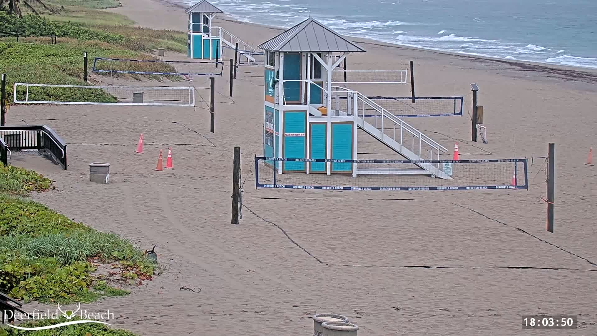 Several volleyball courts are set up on a sandy beach under an overcast sky, with lifeguard stands nearby and a calm ocean in the background.