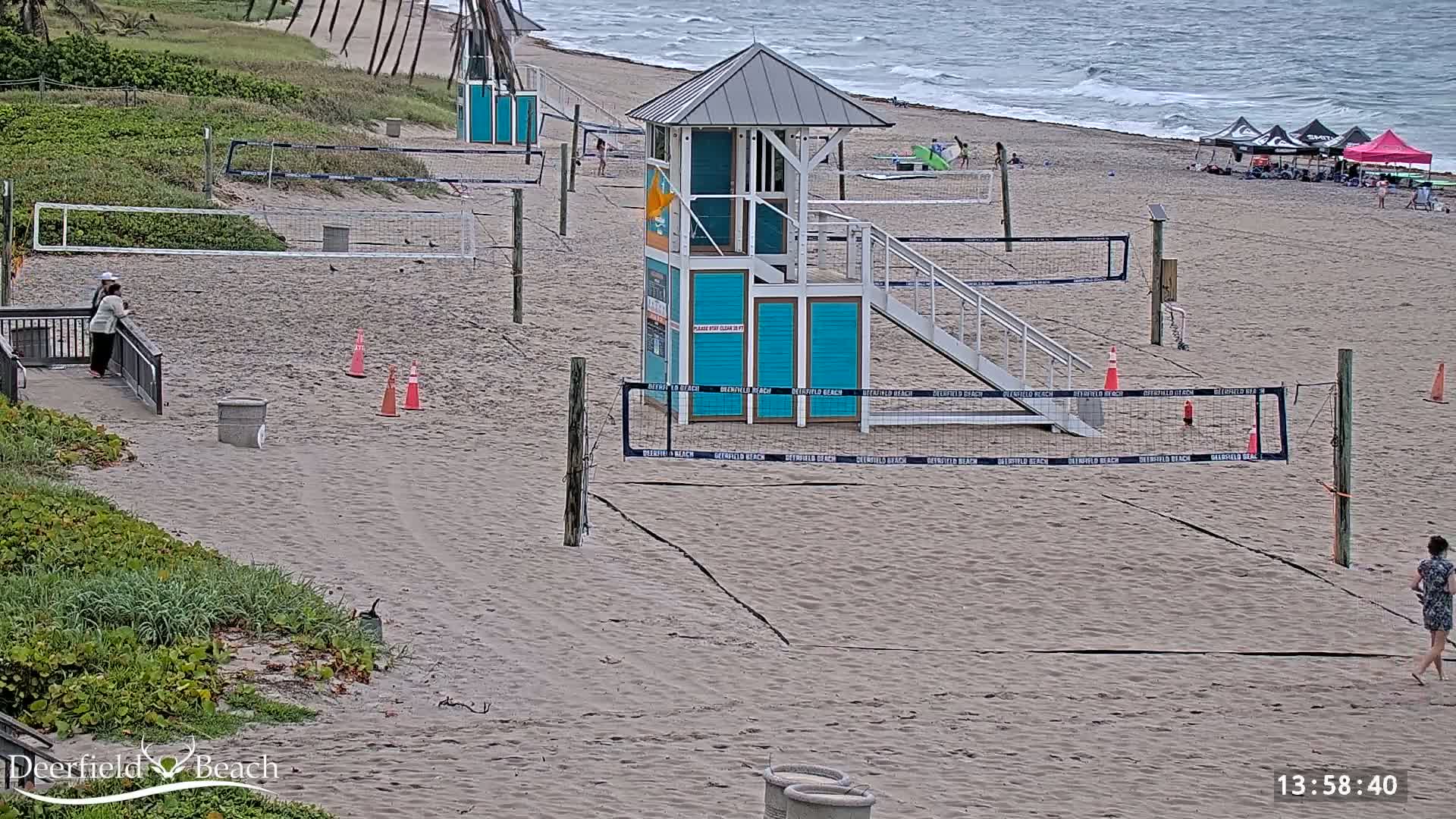 A sandy beach with several volleyball courts, a lifeguard stand, and a few people; the ocean is visible in the background under overcast skies.
