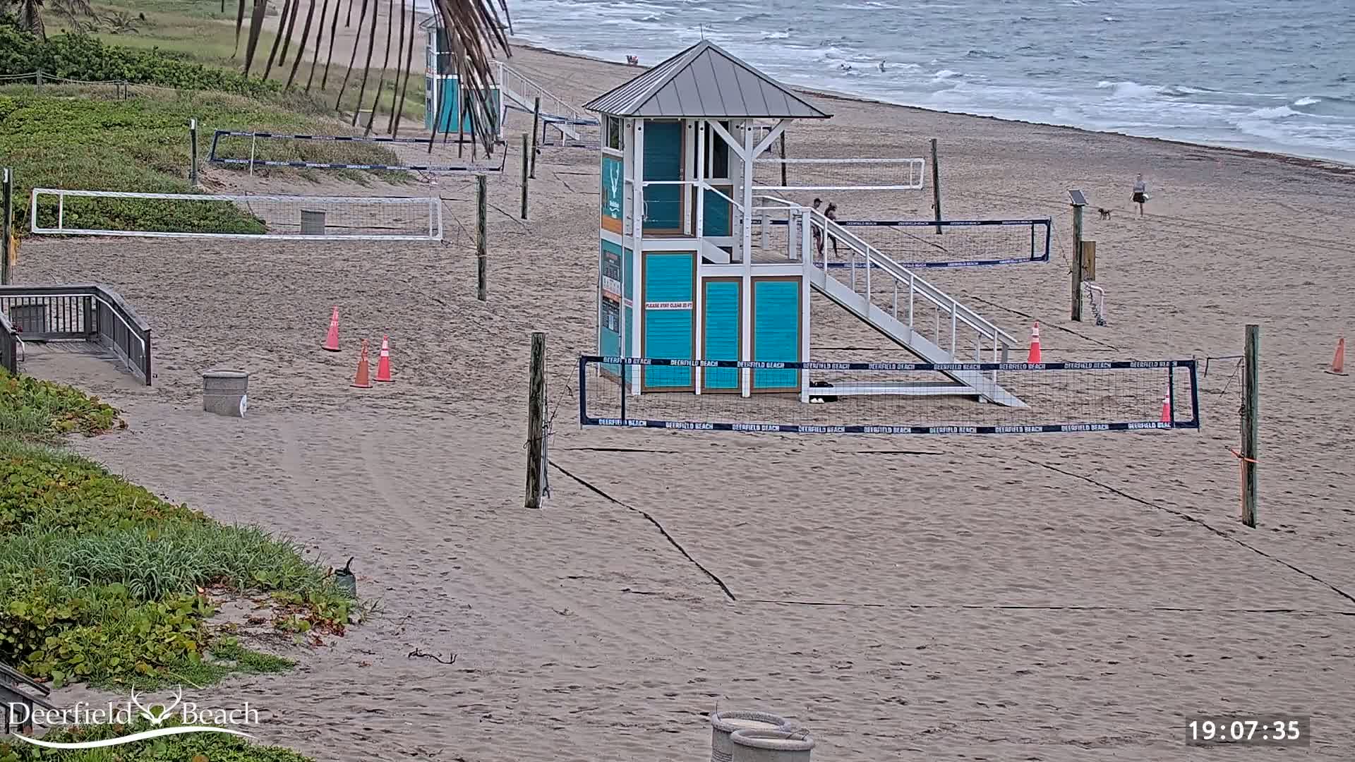 A sandy beach with several volleyball courts, a lifeguard stand, and a calm ocean under an overcast sky.
