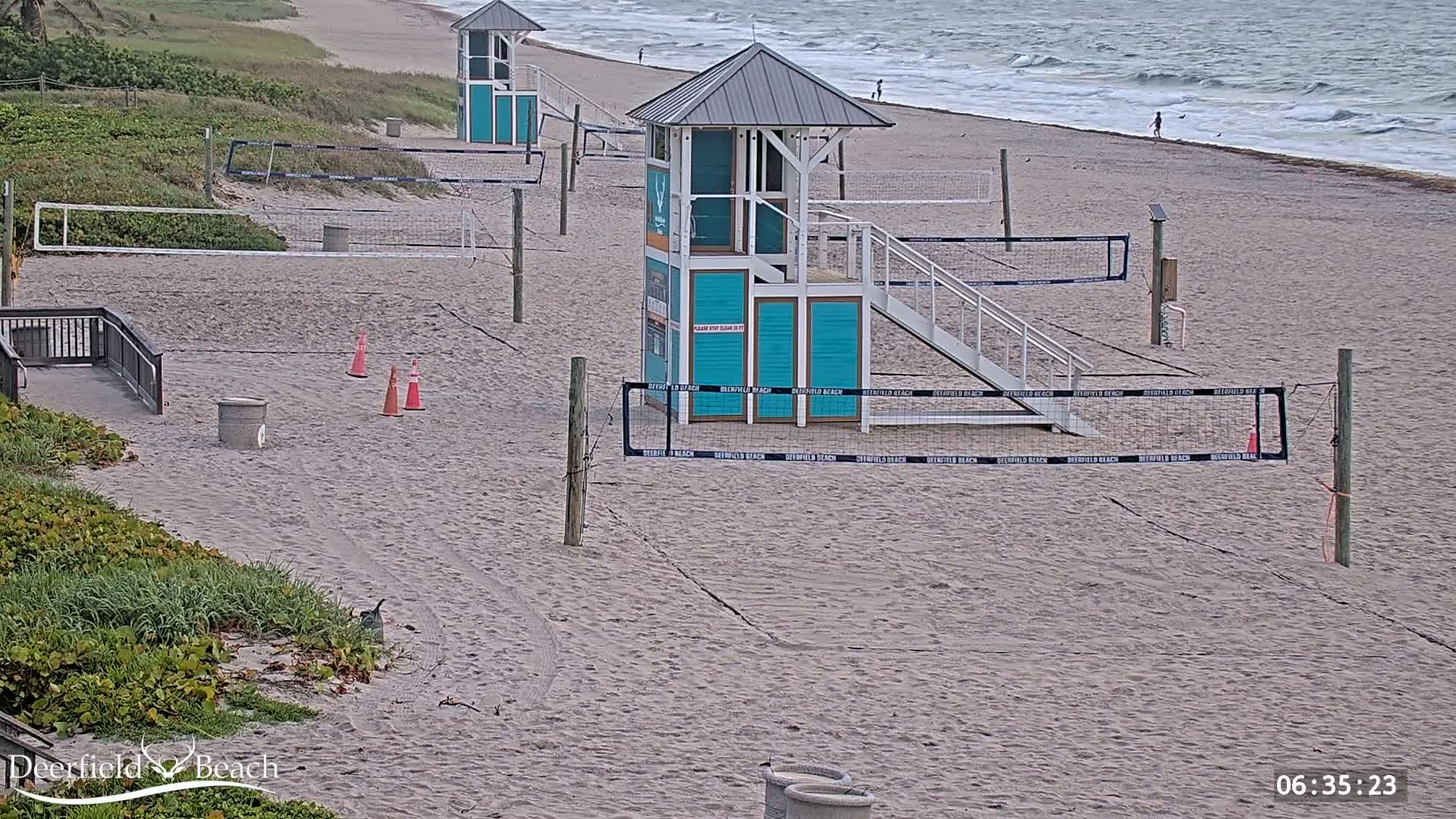 Several volleyball courts are set up on a sandy beach next to a calm ocean under an overcast sky.