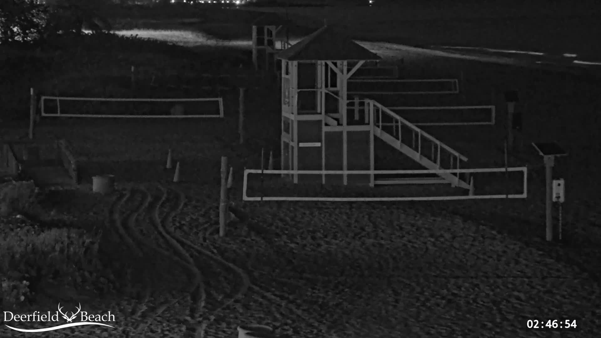 A dark, nighttime view of a sandy beach reveals a prominent lifeguard tower, two volleyball nets, vehicle tracks, and distant lights along the shoreline.