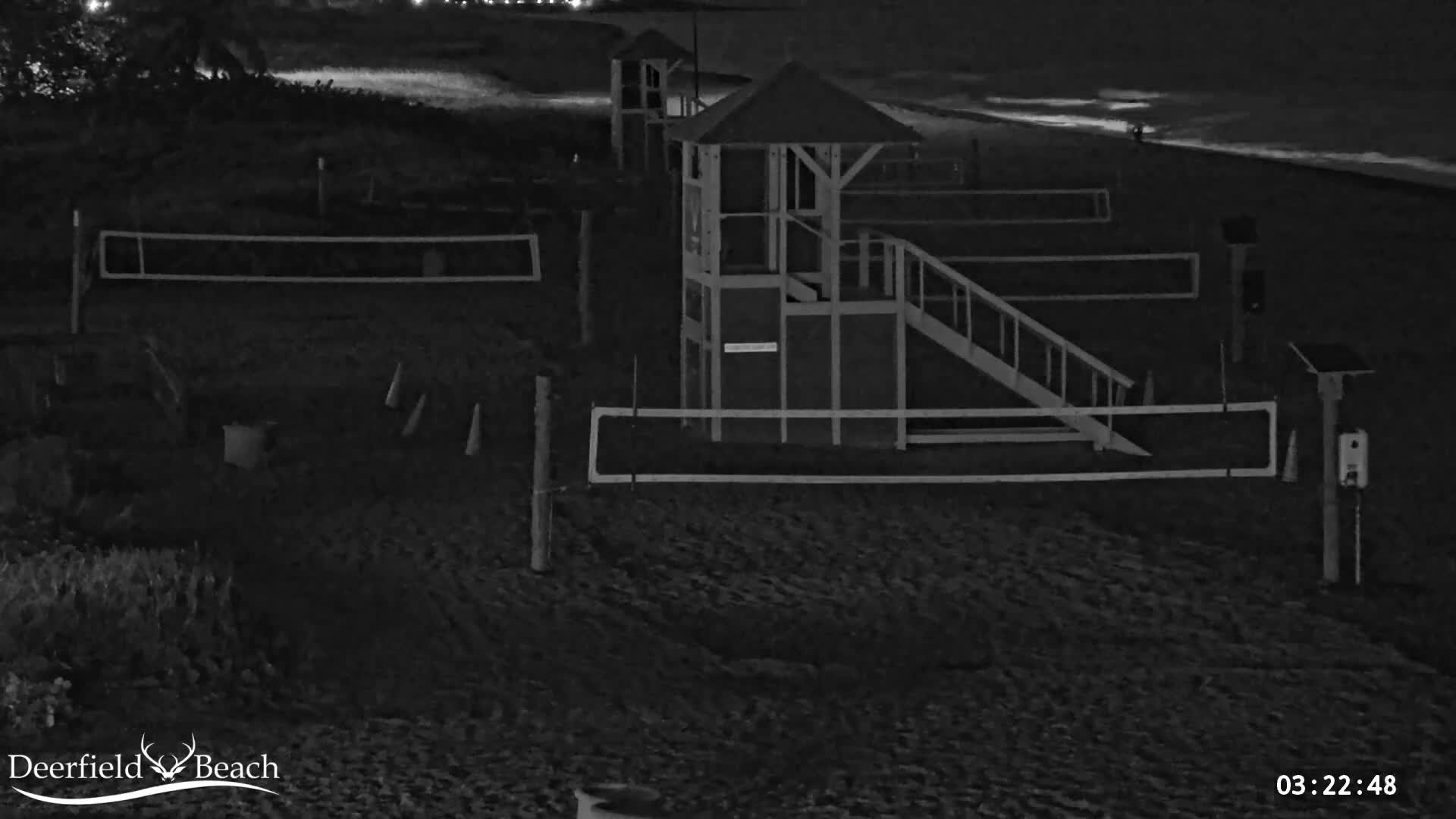 A dark, clear night scene on a calm beach features multiple volleyball court outlines, two elevated wooden structures resembling lifeguard stands, and gentle waves along the distant shoreline.